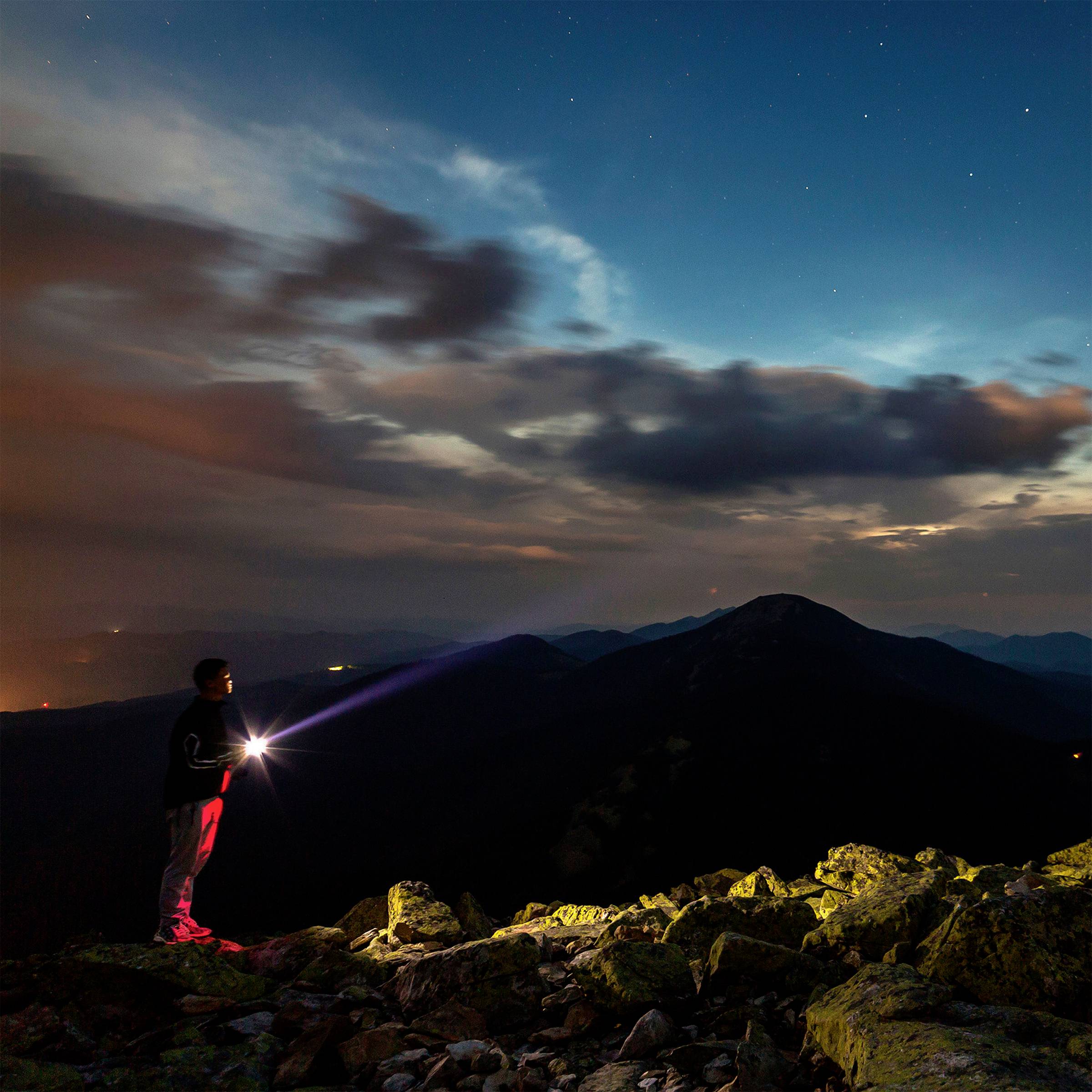Eine Person steht nachts mit einer Taschenlampe auf einem Berggipfel. Der Himmel zeigt Wolken und Sterne, und entfernte Lichter leuchten.