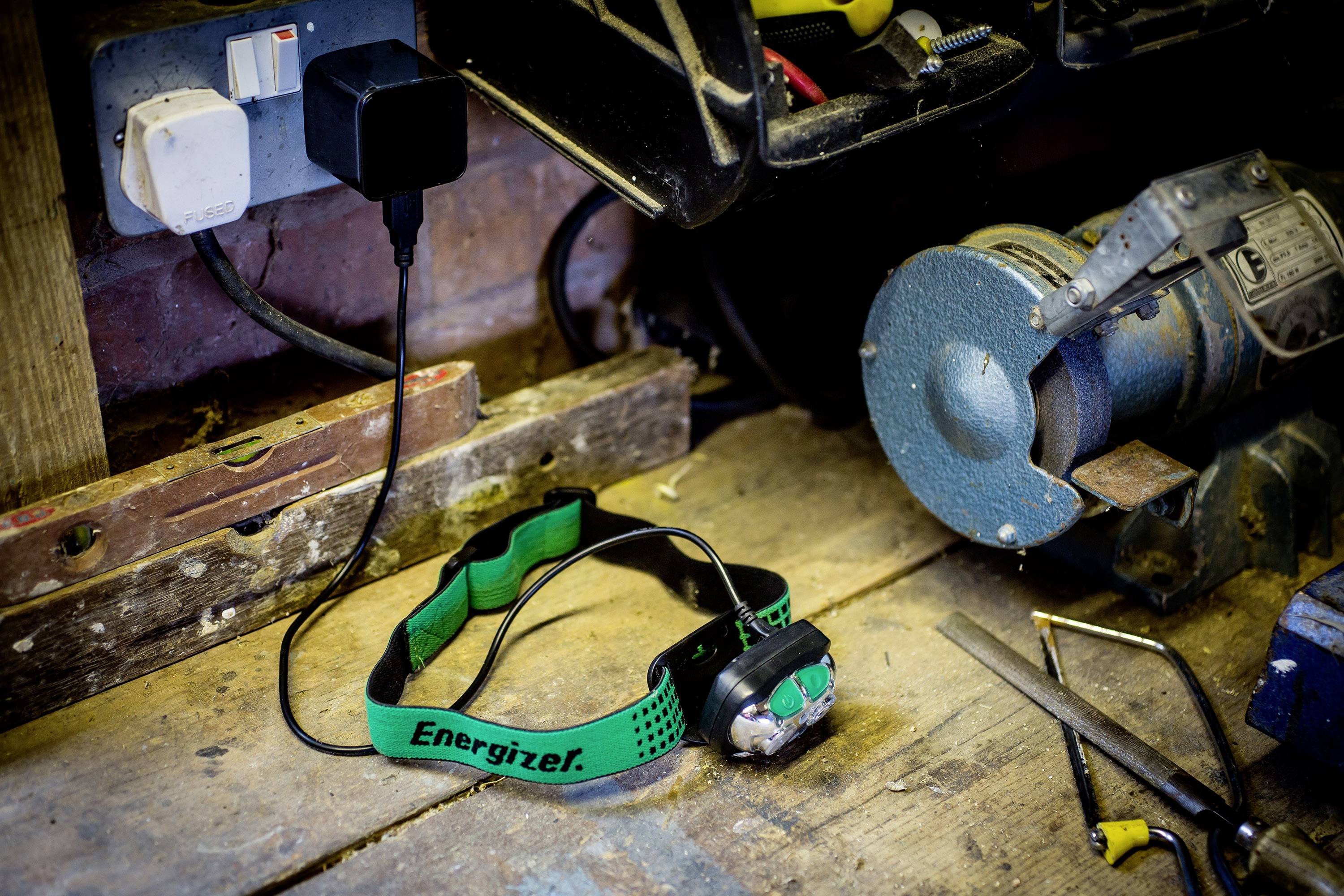 'Arbeitswerkbank mit angeschlossenem Ladegerät, Stirnlampe mit 'Energizer'-Aufdruck, Schleifmaschine und Werkzeugen, die im Hintergrund liegen.'