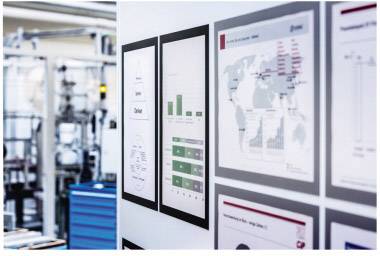 Zwei Reihen von Schaubildern und Diagrammen auf einer Wand. Links ist eine unscharfe, industrielle Umgebung erkennbar.