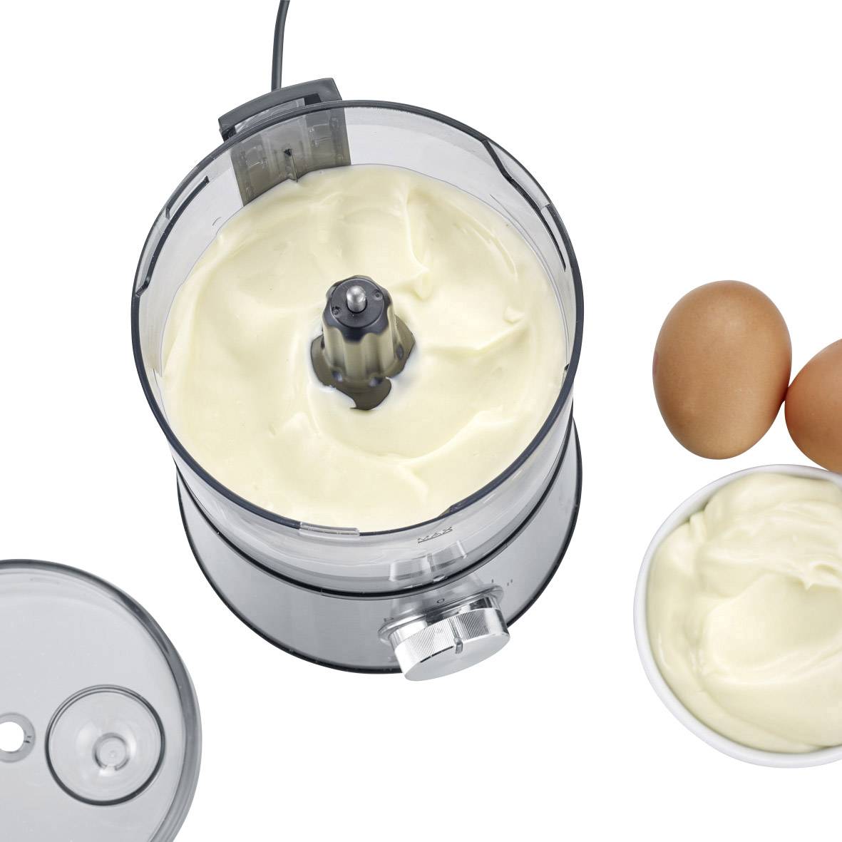 Ein Mixer mit weißer Creme, daneben zwei Eier und eine Schale mit ähnlicher Creme auf einer weißen Oberfläche.