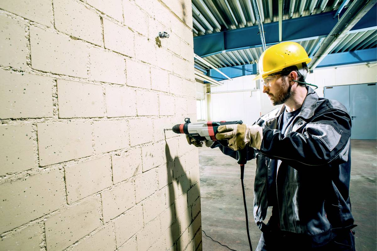 Ein Arbeiter trägt Schutzkleidung und einen Helm, während er mit einer Bohrmaschine ein Loch in eine Ziegelwand bohrt.