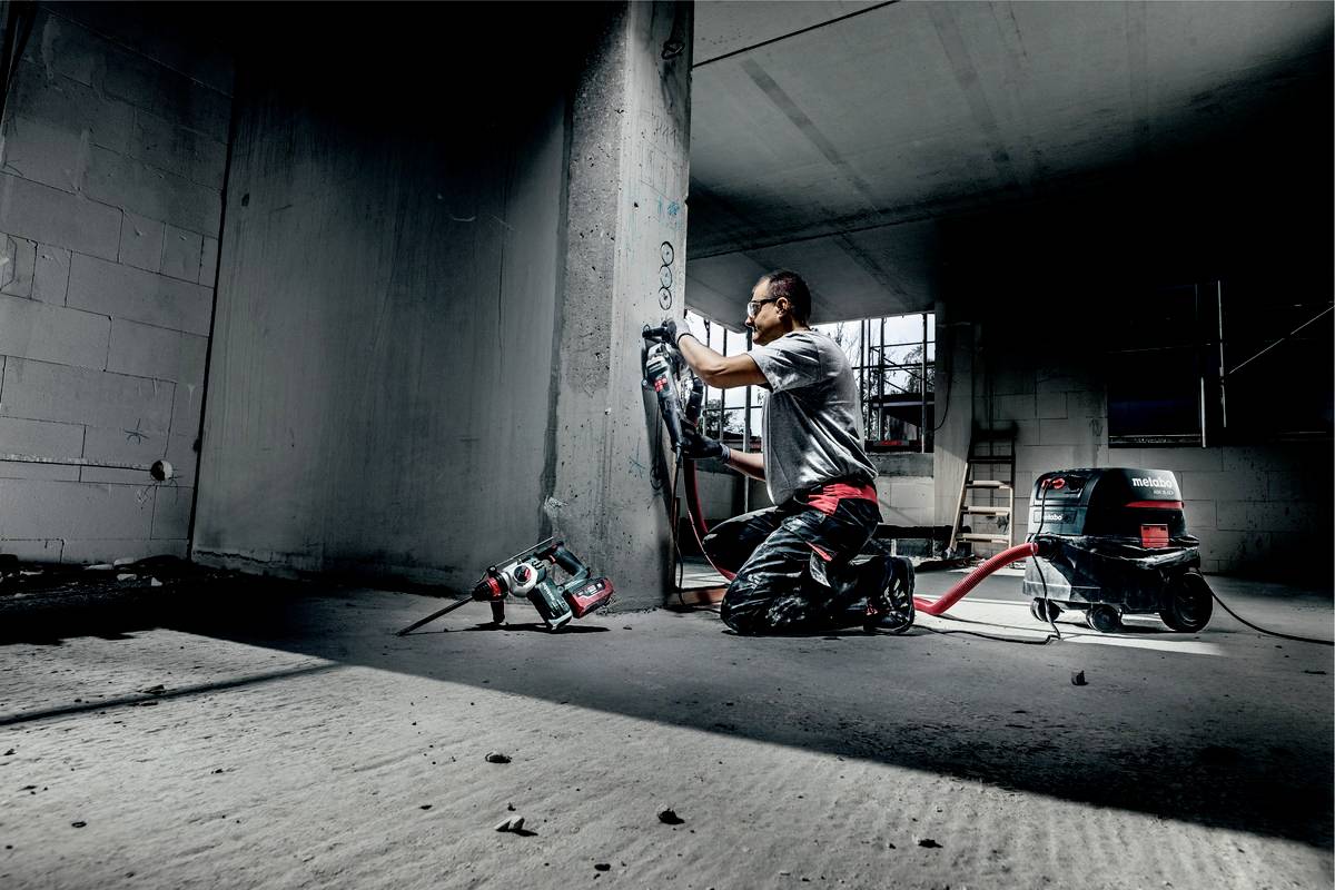 Ein Bauarbeiter kniet und arbeitet mit einem Bohrhammer an einer Betonwand in einem halbfertigen Gebäude. Eine Staubabsaugung ist angeschlossen.