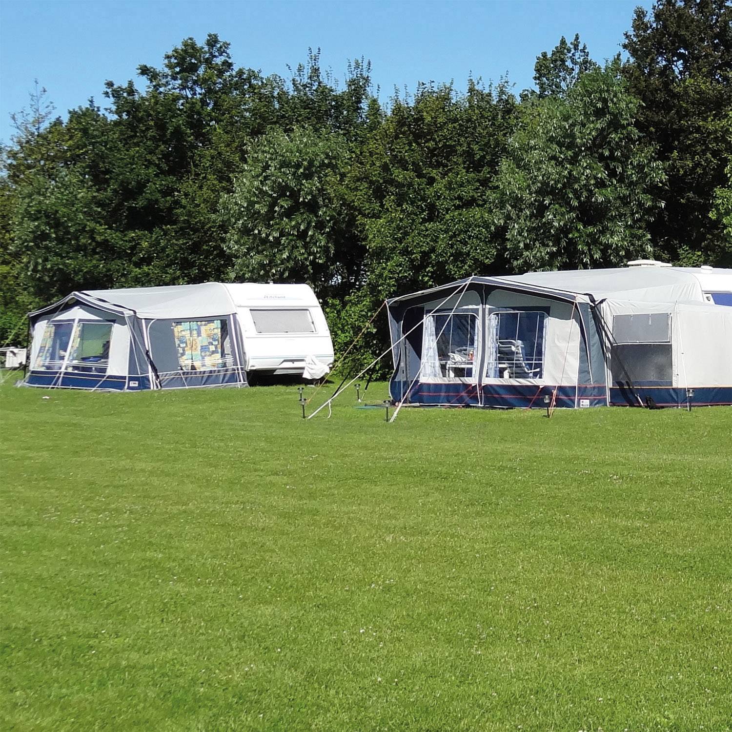 Zwei große Campingzelte auf einer grünen Wiese, umgeben von Bäumen. Sonniges Wetter, klare blaue Himmel mit vereinzelt Wolken.
