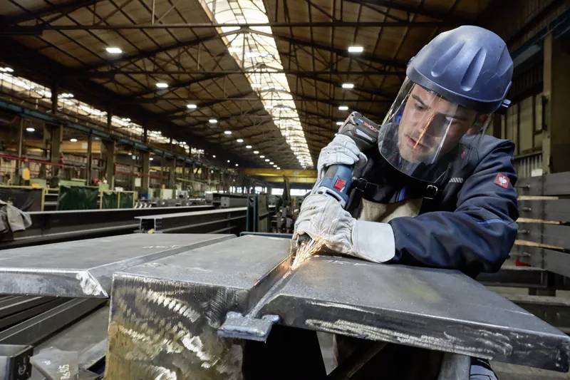 Ein Arbeiter in Schutzausrüstung verwendet ein Elektrowerkzeug zum Schneiden von Metall in einem Industrielager, umgeben von Stahlträgern und hellem Licht.