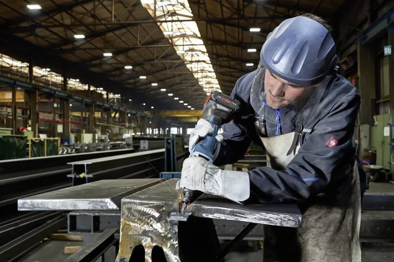 Ein Arbeiter in Schutzausrüstung verwendet ein elektrisches Werkzeug, um an einem großen Metallstück in einer industriellen Lagerhalle zu arbeiten.