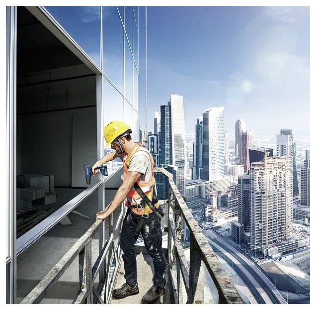 Ein Bauarbeiter mit Sicherheitsausrüstung installiert ein Fenster an einem Hochhaus, wobei eine Stadtsilhouette im Hintergrund zu sehen ist.