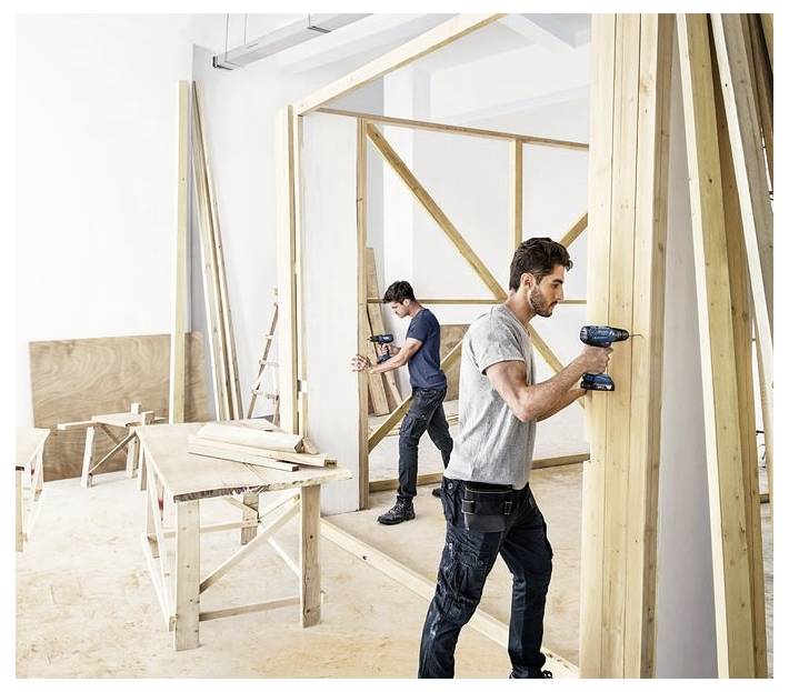 Zwei Personen benutzen Bohrmaschinen, um einen Holzrahmen in einer Werkstatt zusammenzubauen, umgeben von verschiedenen Holzstücken und Werkzeugen.