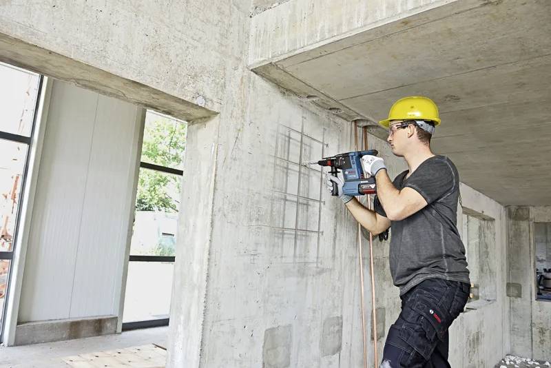 Ein Bauarbeiter mit Schutzhelm verwendet eine Bohrmaschine an einer Betonwand in einem Gebäude im Bau.