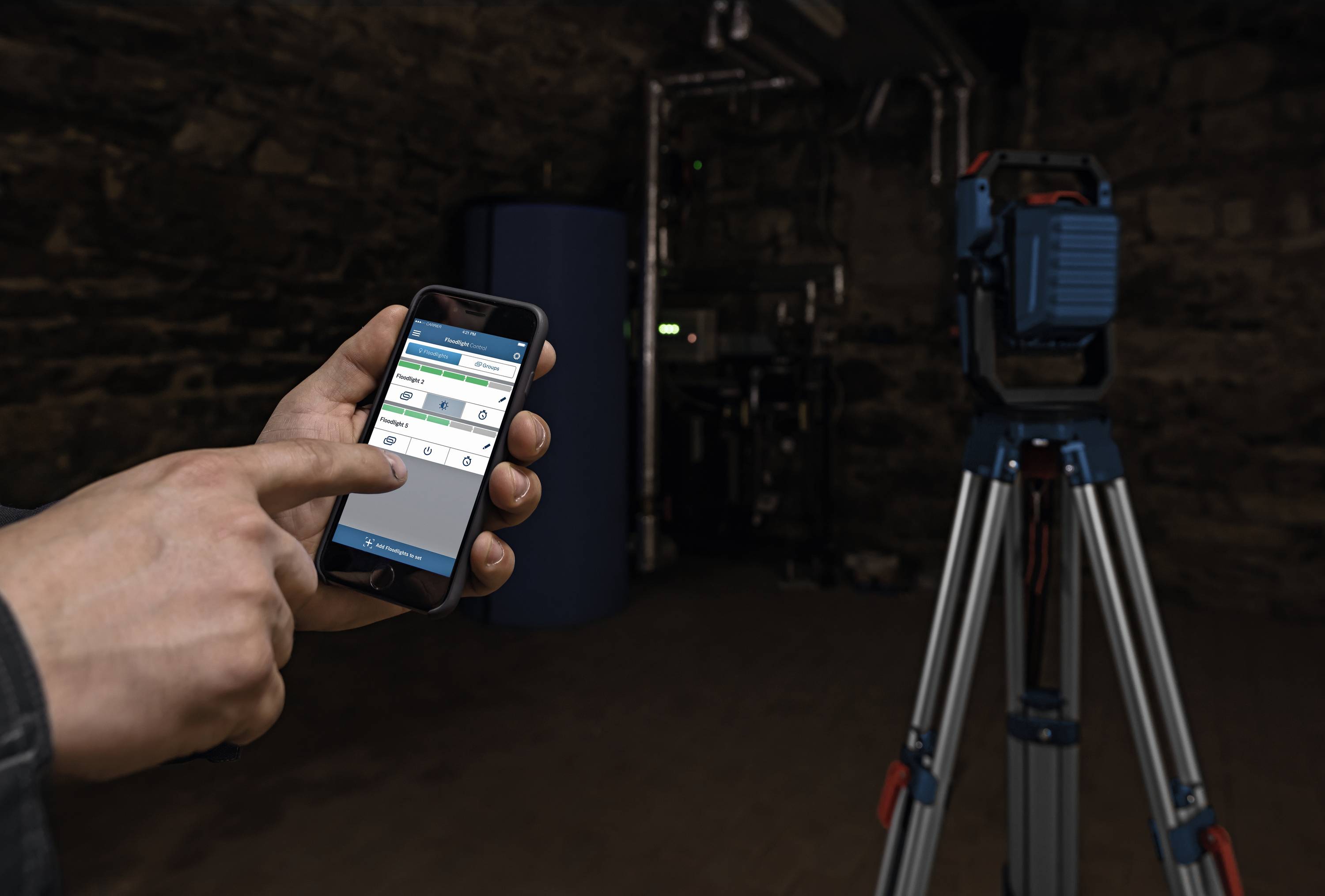 Eine Person bedient eine App auf einem Smartphone zur Steuerung eines Geräts auf einem Stativ in einem industriellen Raum.
