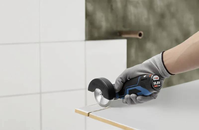 Eine Person mit einem Handschuh verwendet ein kompaktes Elektrowerkzeug, um eine Kachel zu schneiden. Das Werkzeug ist schwarz und blau, und die Umgebung scheint ein Arbeitsbereich zu sein.