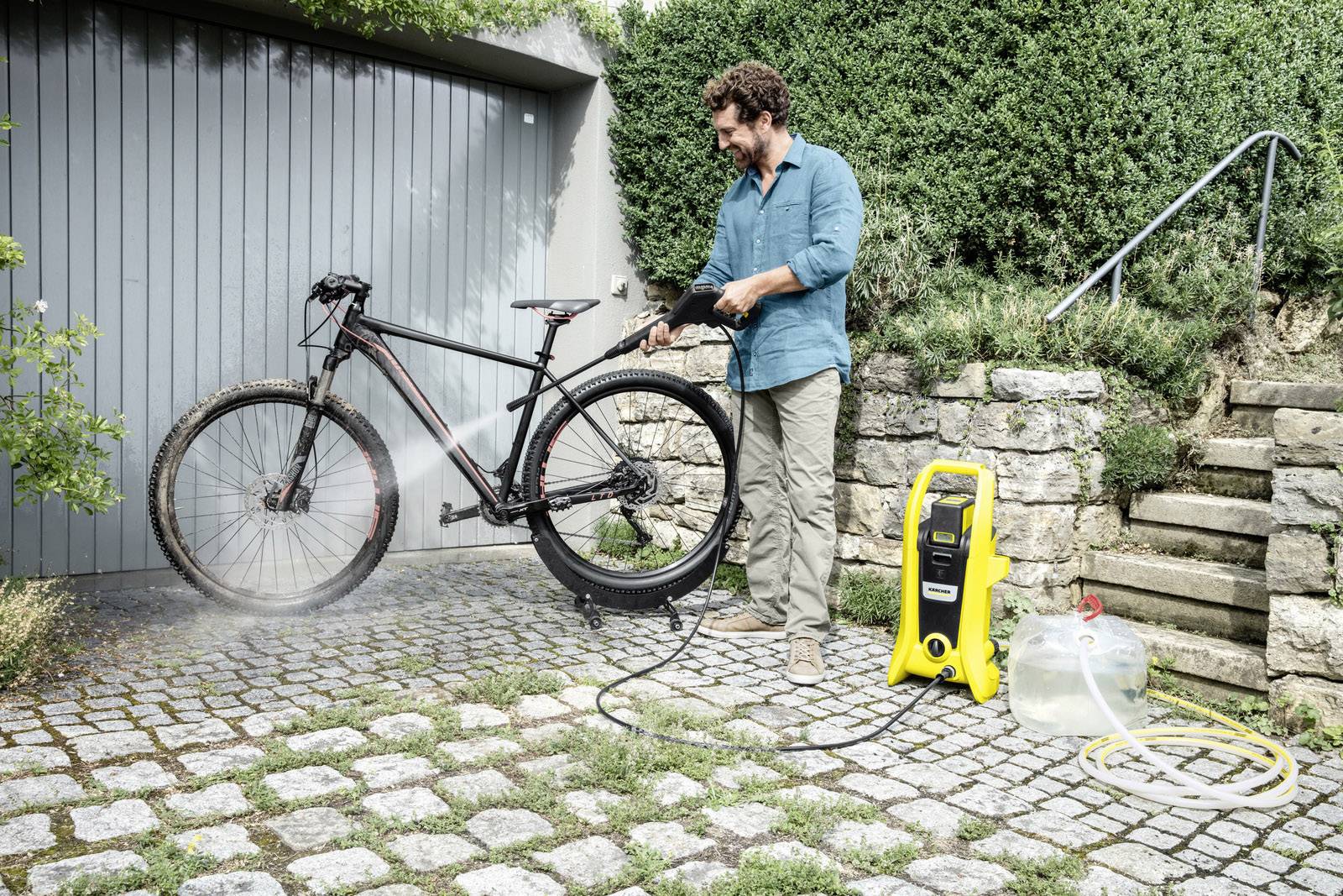 Ein Mann reinigt ein schwarzes Fahrrad mit einem Hochdruckreiniger auf einer gepflasterten Fläche, mit einer Hecke im Hintergrund.