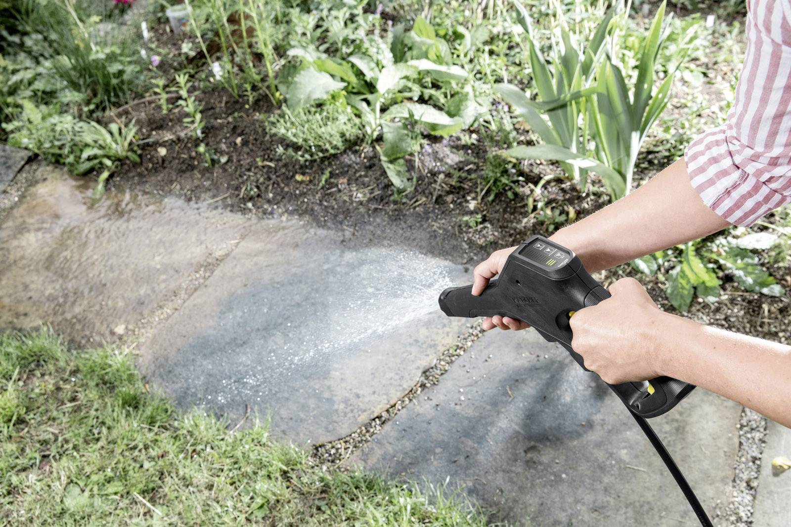 Eine Person reinigt mit einem Hochdruckreiniger eine Gartenplatte. Umgeben von Gras und Pflanzen.