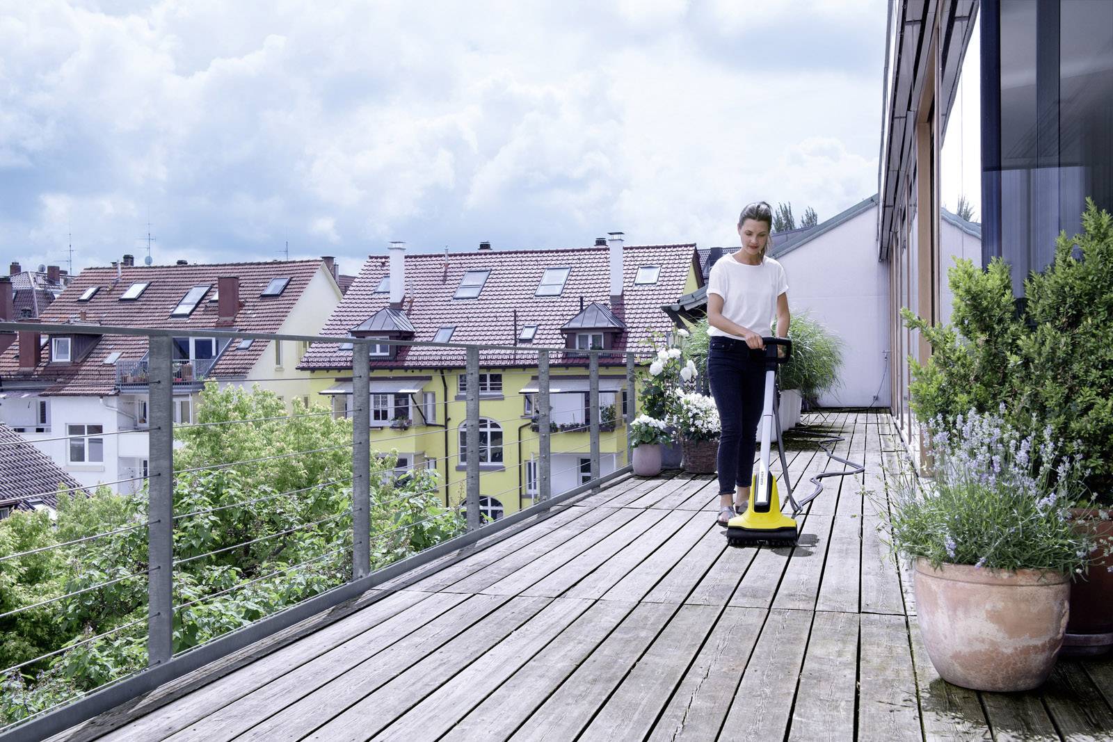 Eine Person reinigt eine Holzterrasse mit einem gelben Hochdruckreiniger. Im Hintergrund sind Wohnhäuser und Bäume zu sehen.