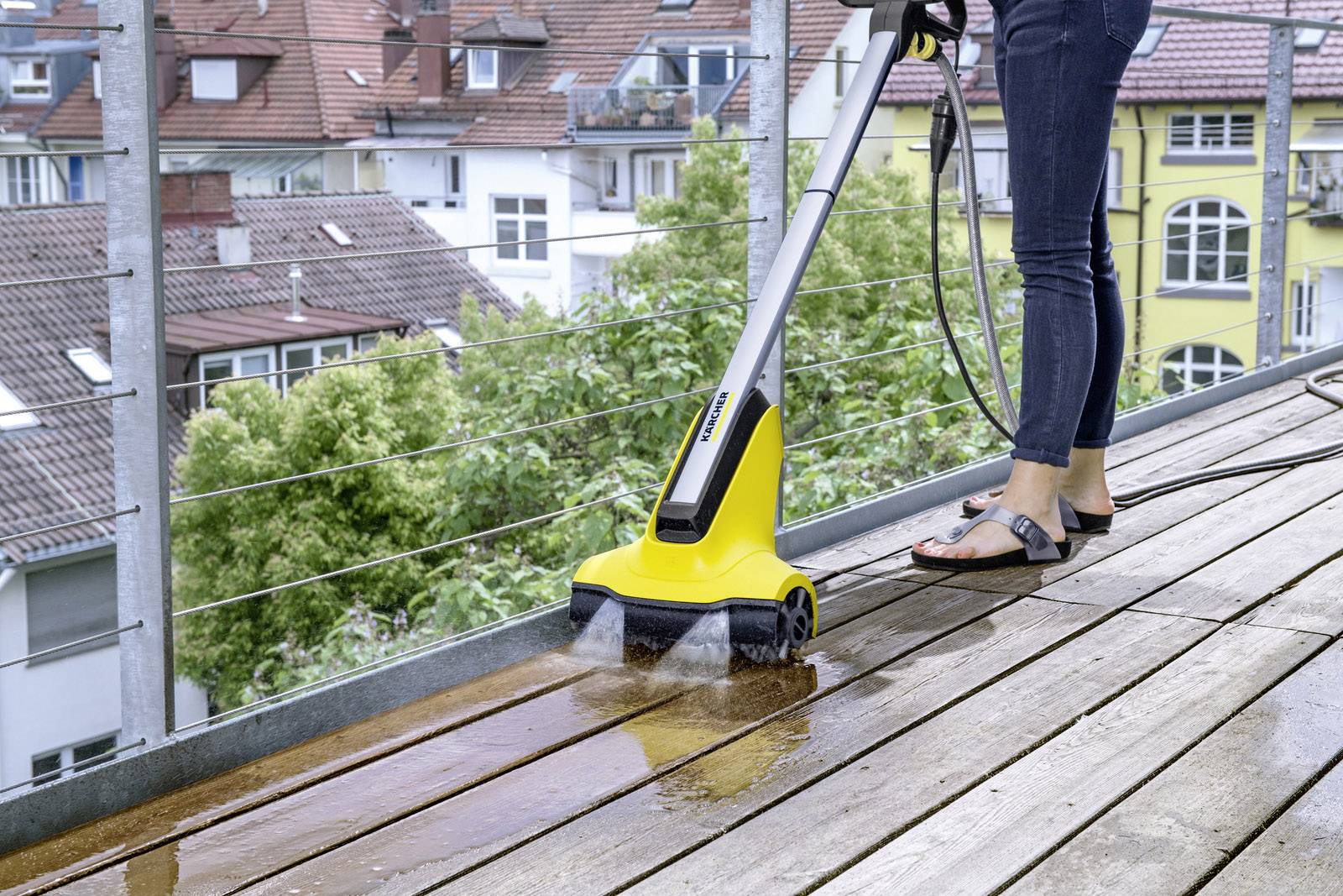 Person mit gelber Reinigungsmaschine säubert Holzboden auf Balkon; im Hintergrund sind Häuser und Bäume zu sehen.