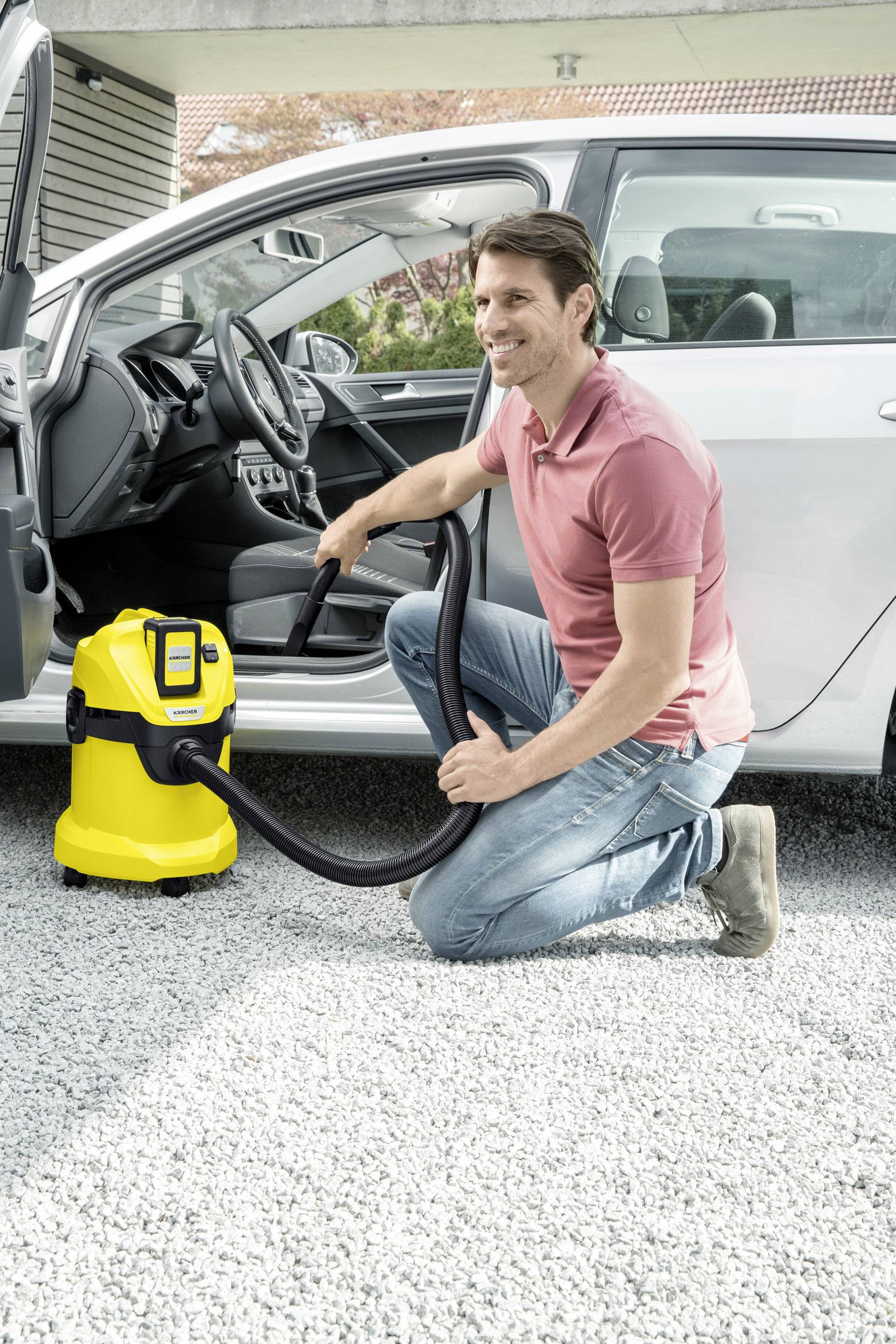 Ein Mann kniet neben einem silbernen Auto und benutzt einen gelben Industriestaubsauger, um den Innenraum zu reinigen.