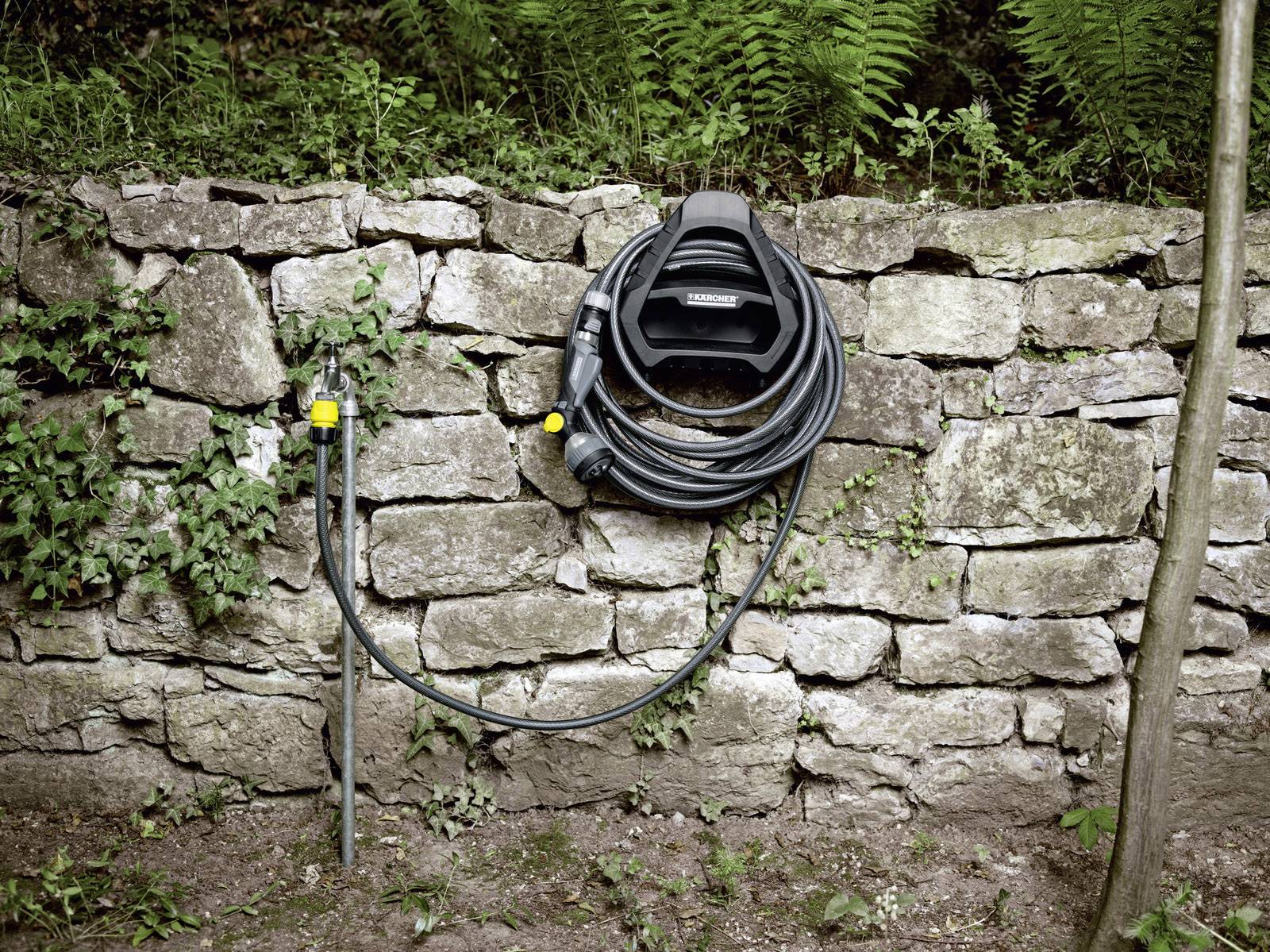 Gartenschlauch an der Wand eines Steingartens befestigt, umgeben von grünen Farnen und Pflanzen. Links ein Wasserhahn mit gelbem Anschluss.