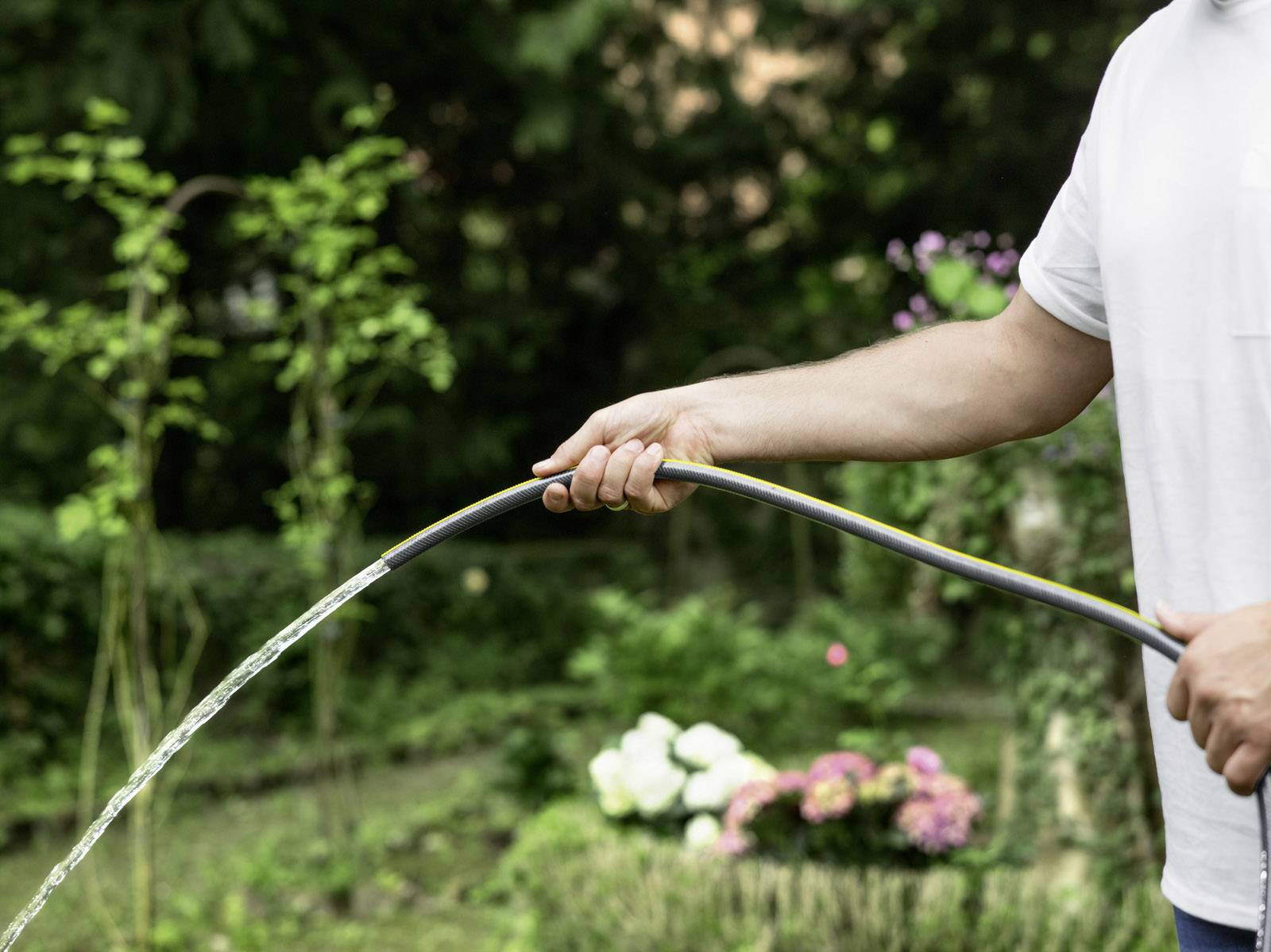 Person gießt Pflanzen im Garten mit Schlauch. Hintergrund hat blühende Blumen und grüne Sträucher.