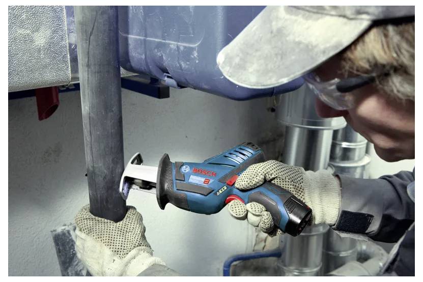 Eine Person verwendet eine kabellose Säbelsäge, um ein Rohr zu durchtrennen; sie trägt Sicherheitsbrille und Handschuhe, was auf eine Arbeitsumgebung hindeutet.