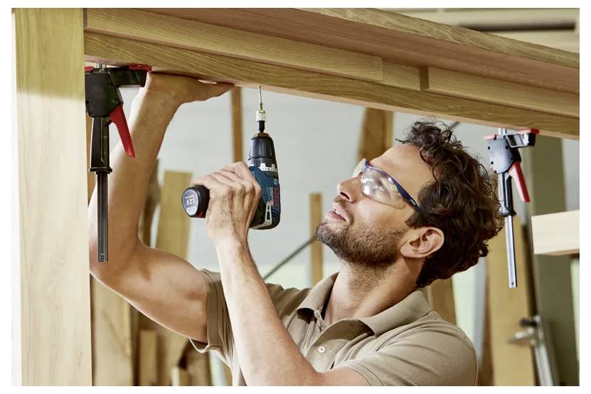 Ein Mann mit Schutzbrille benutzt eine Elektrobohrmaschine, um einen Holzrahmen zusammenzubauen und arbeitet dabei über Kopfhöhe in einer Werkstatt.