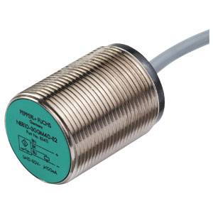Ein zylindrischer, metallischer Sensor mit grünem Etikett zeigt Markierung 'Pepperl+Fuchs'. Ein graues Kabel ist daran angeschlossen.