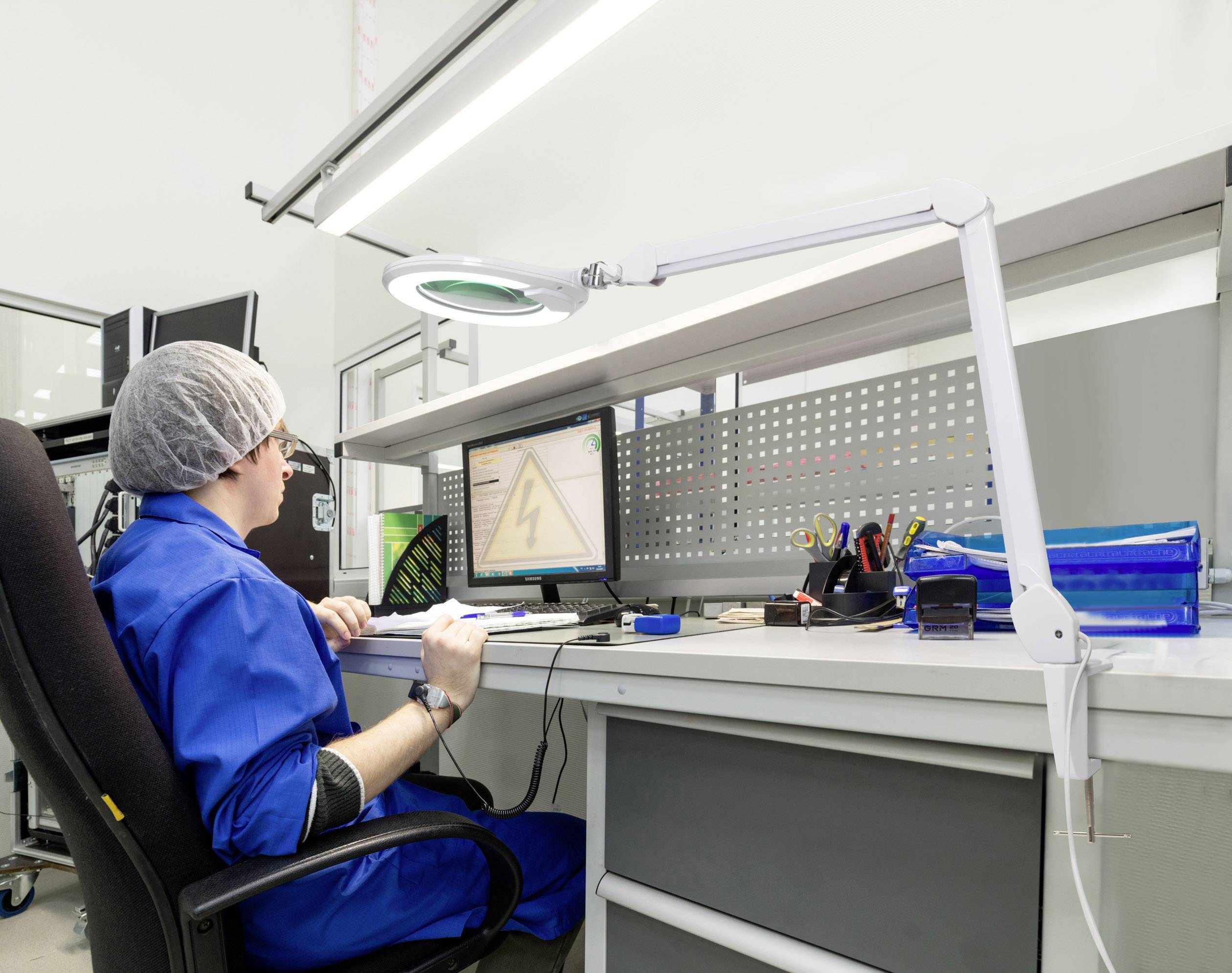 Eine Person in blauer Schutzkleidung und Haarnetz arbeitet am Computer in einer Laborumgebung. Schreibtisch mit Werkzeugen und Monitor.