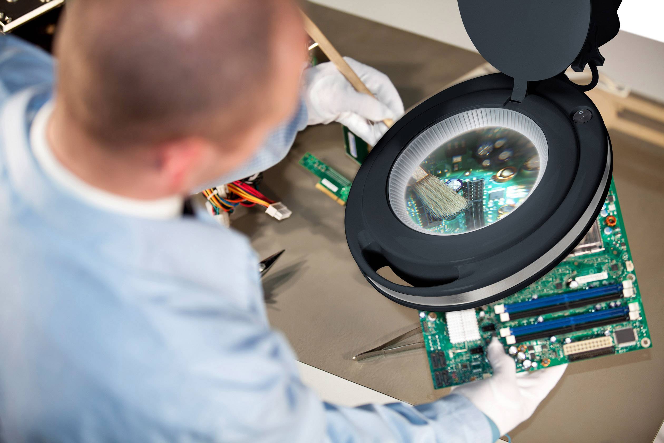 Ein Techniker reinigt eine offene Computerplatine mit einem Pinsel unter einer beleuchteten Lupe auf einem Tisch.