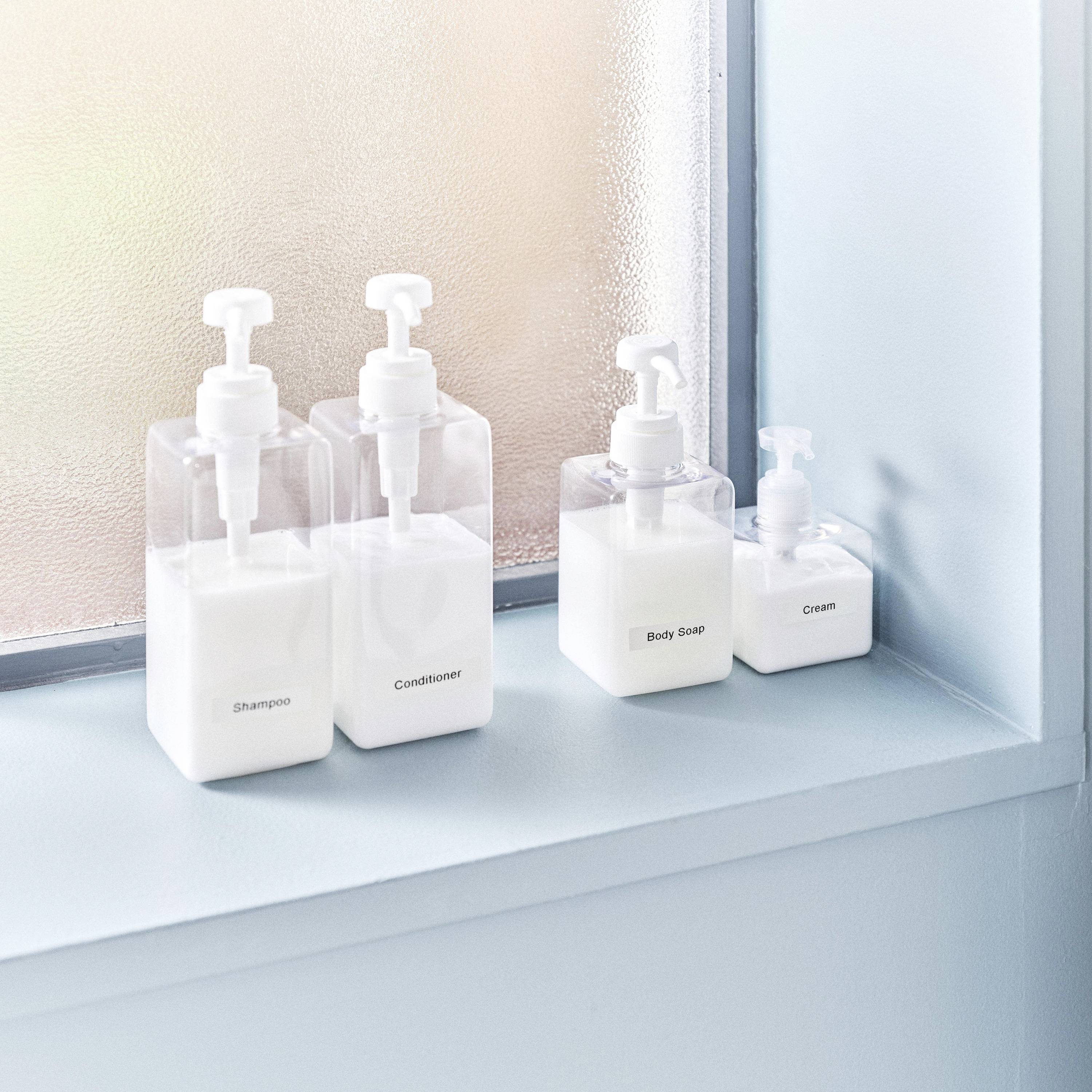Vier Spenderflaschen mit 'Shampoo', 'Conditioner', 'Body Soap' und 'Body Cream' stehen auf einem Fensterbrett aus Glas.