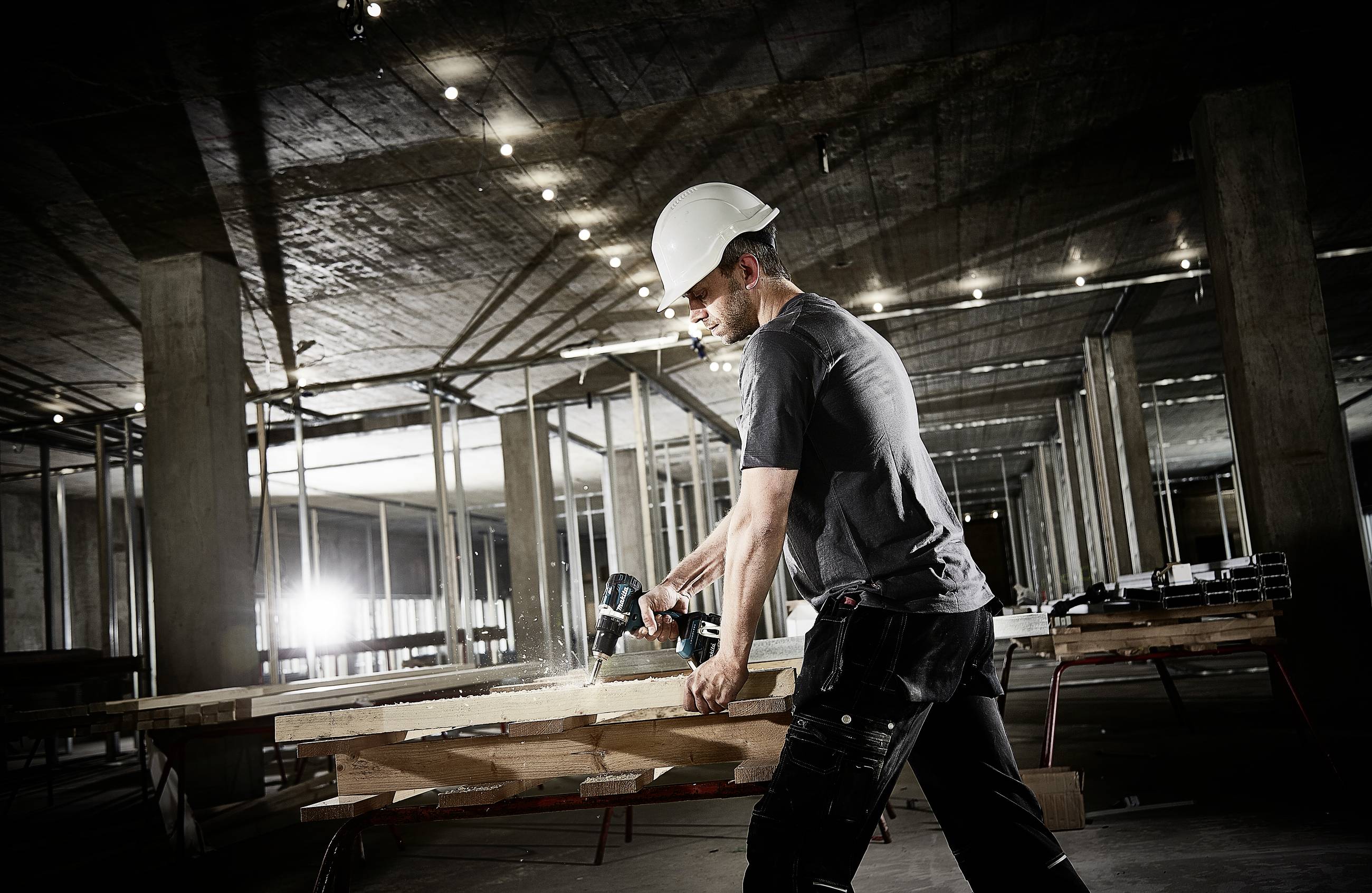 Ein Bauarbeiter mit Schutzhelm sägt Holz auf einer Baustelle. Der Hintergrund zeigt unvollständige Betonstrukturen und schwaches Licht.