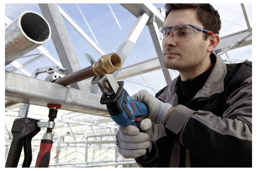 Eine Person mit Sicherheitsbrille und Handschuhen verwendet eine Säbelsäge, um ein Metallrohr in einer leichten Industrieumgebung zu schneiden.