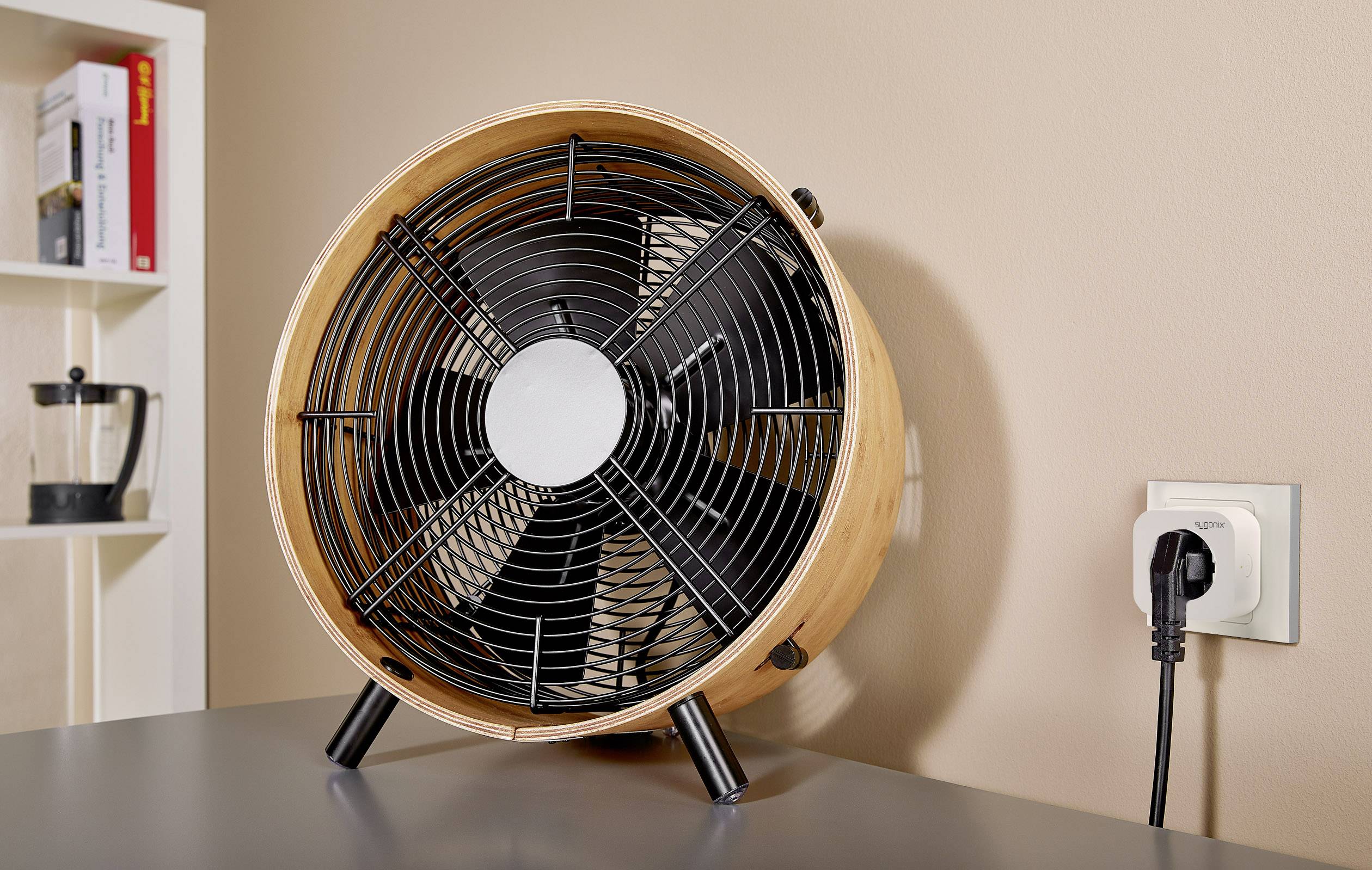 Ein runder Tischventilator in Holzoptik steht auf einem Tisch. Rechts daneben ist eine Steckdose mit einem eingesteckten Stecker. Im Hintergrund ist ein Bücherregal erkennbar.