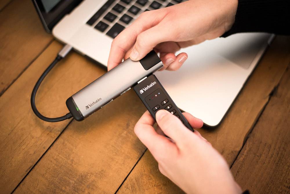 Person hält einen Verbatim USB-Stick und einen drahtlosen Präsentationsklicker über einem Laptop, bereit zur Nutzung.