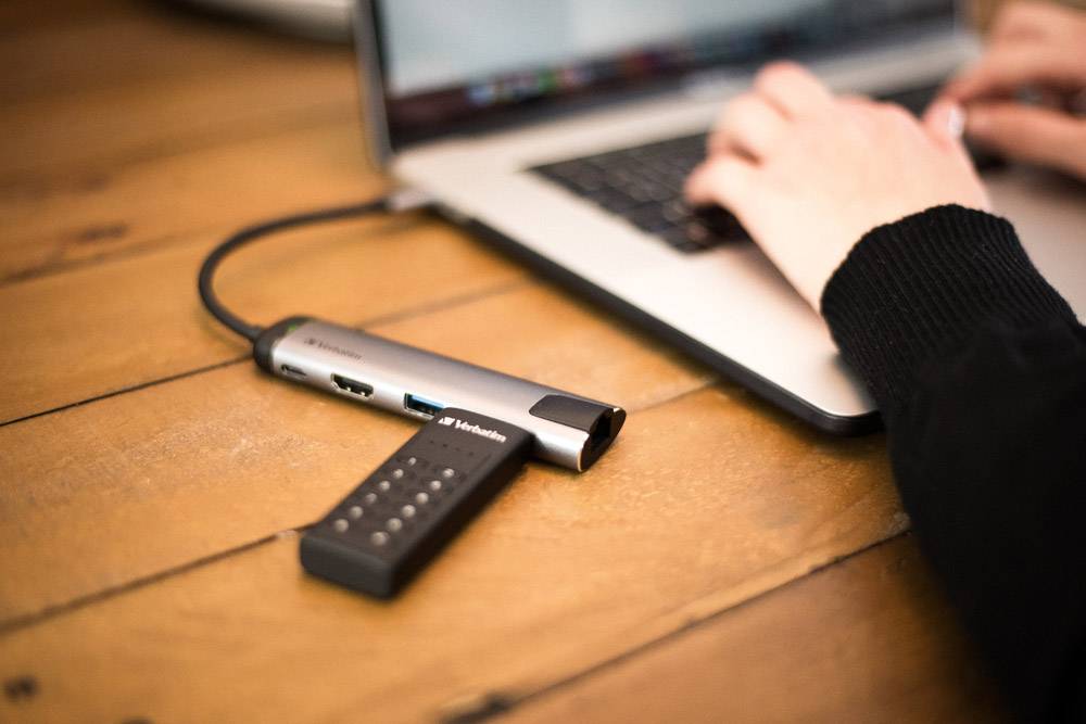 Person arbeitet an einem Laptop, verbunden mit einem USB-Hub auf einem Holztisch.
