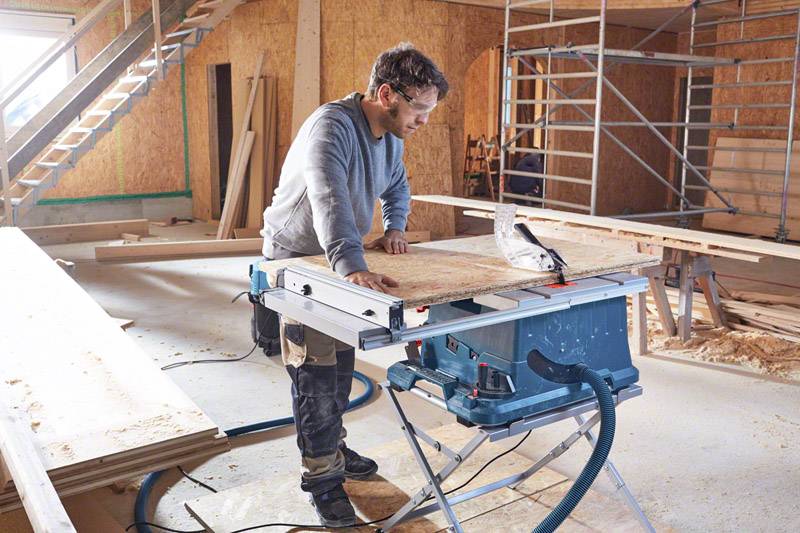 Ein Mann arbeitet in einer Werkstatt mit einer Tischkreissäge und schneidet Holzplatten zu. Im Hintergrund sind Gerüste und Holzmaterialien.