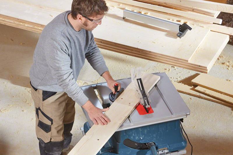 Eine Person sägt ein Holzbrett auf einer Tischsäge, umgeben von Holzspänen. Sie trägt Schutzkleidung und arbeitet konzentriert.