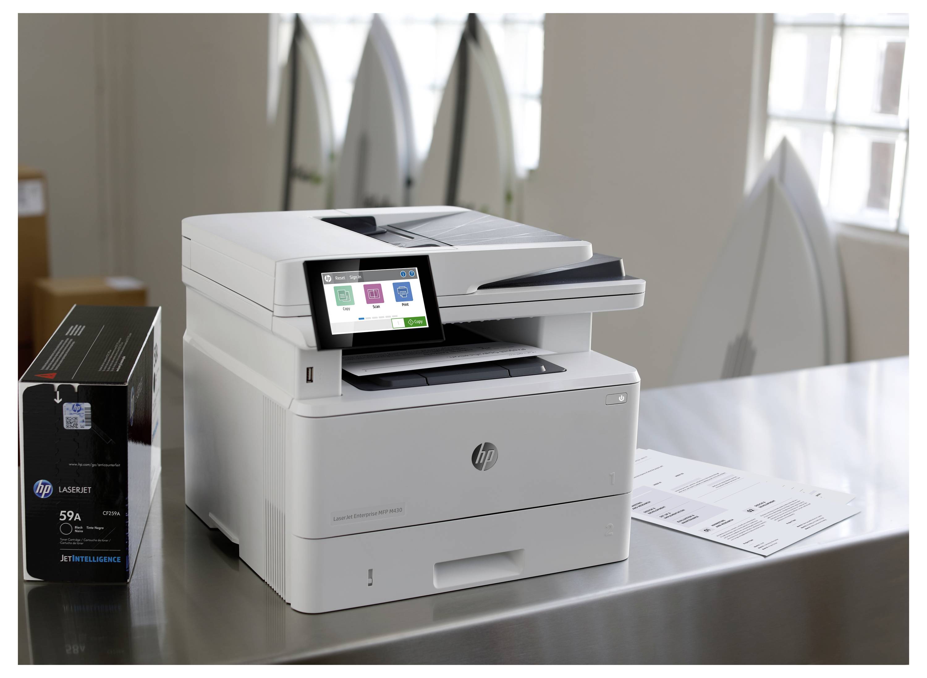 Ein HP-Multifunktionslaserdrucker auf einem Tisch neben einer Tonerbox mit Touchscreen-Display und Papierausgabefach in einer Büroumgebung.