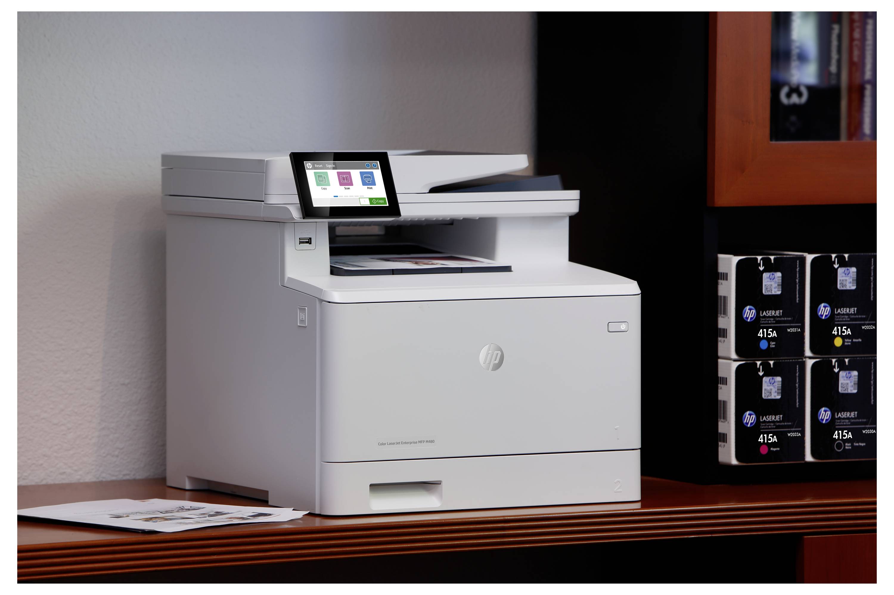 Weißer HP Multifunktionsdrucker/Kopierer auf einem Holztisch neben Tintenpatronen-Boxen, mit sichtbarem Touchscreen-Bedienfeld.