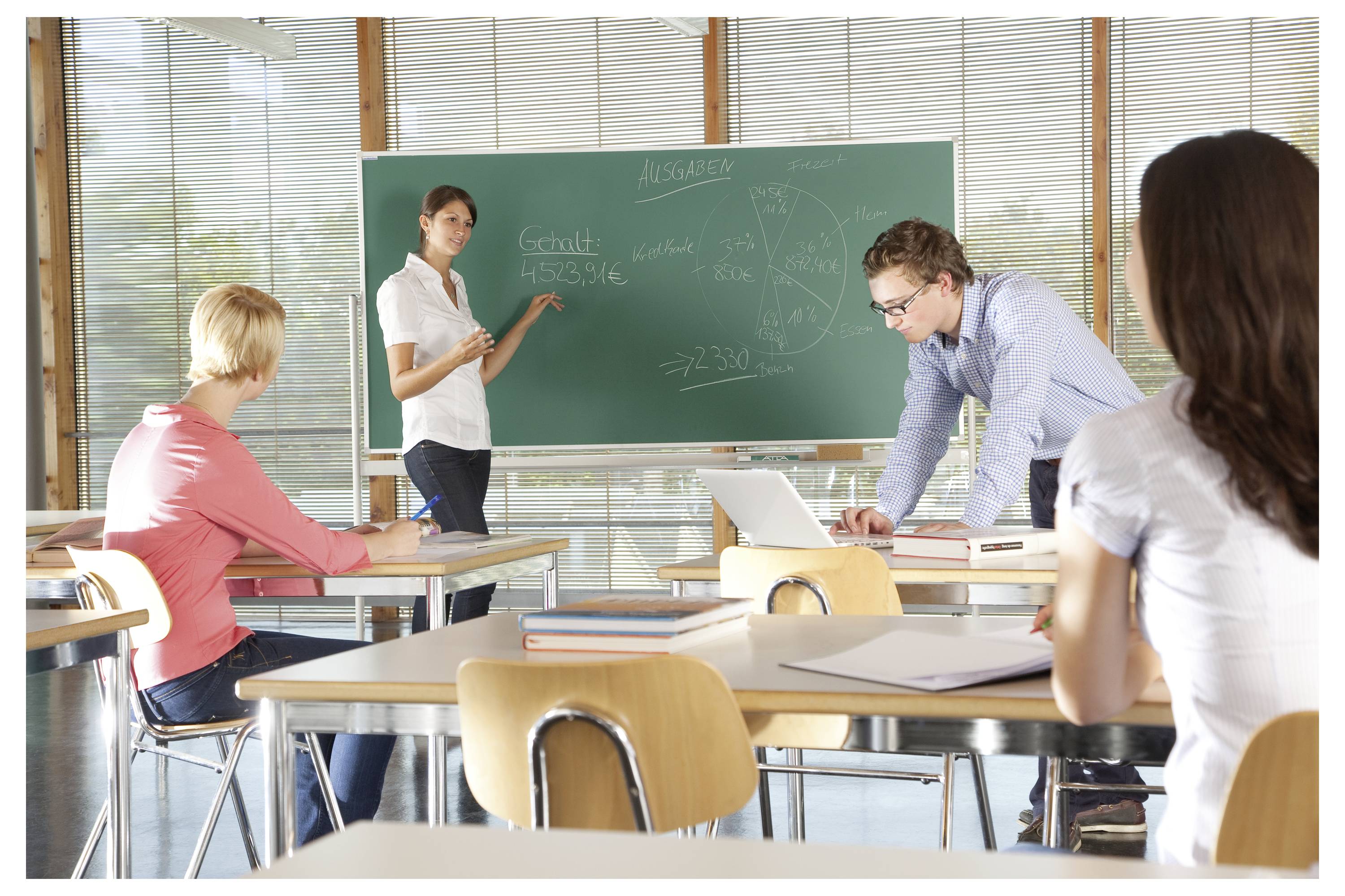 Lehrer präsentiert an einer Tafel, während Schüler an Schreibtischen sitzen und ein Mann an einem Laptop in einem hellen Klassenzimmer arbeitet.