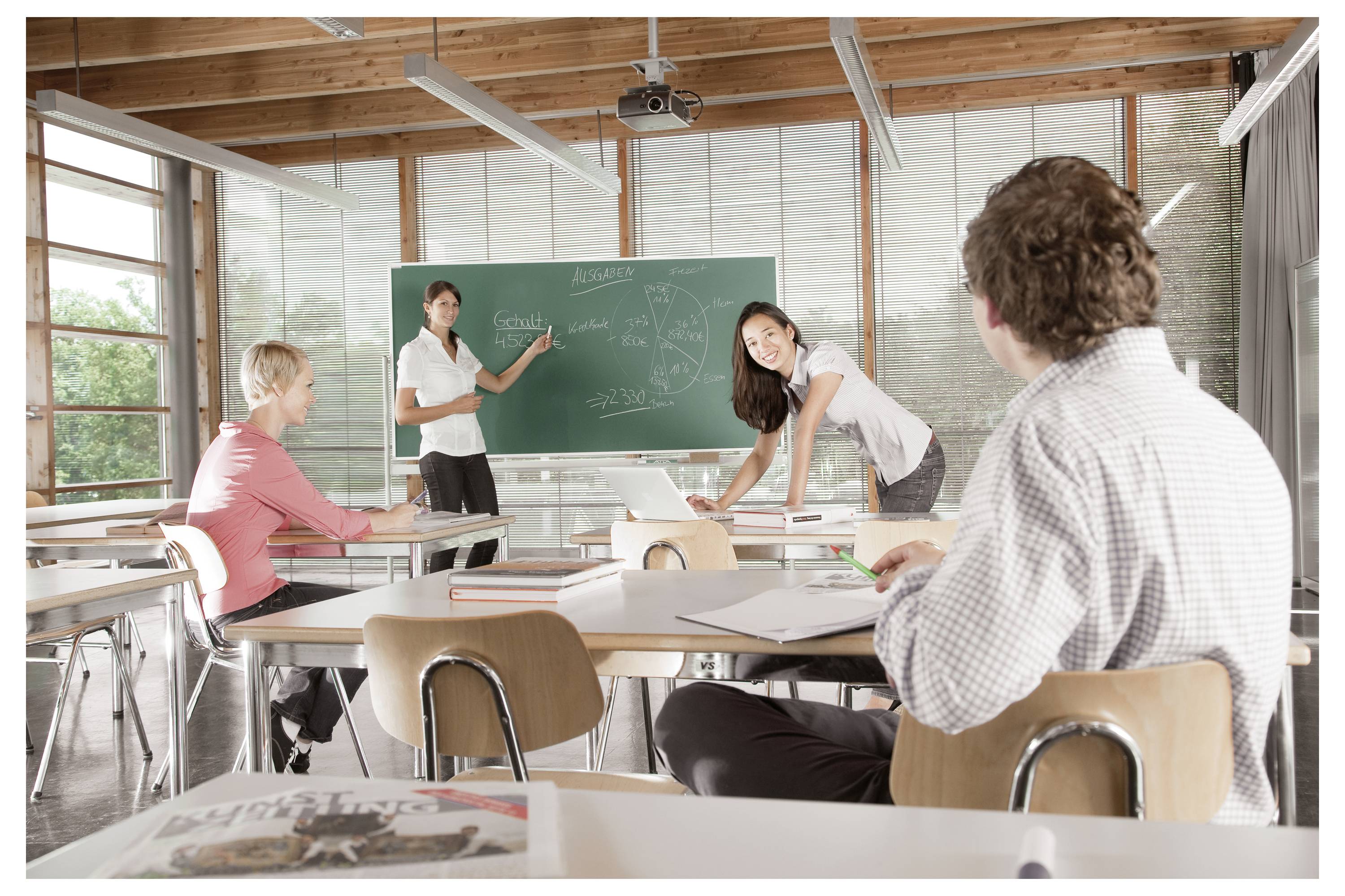 Schüler im Klassenzimmer beobachten einen Lehrer, der während des Unterrichts auf eine Tafel zeigt.