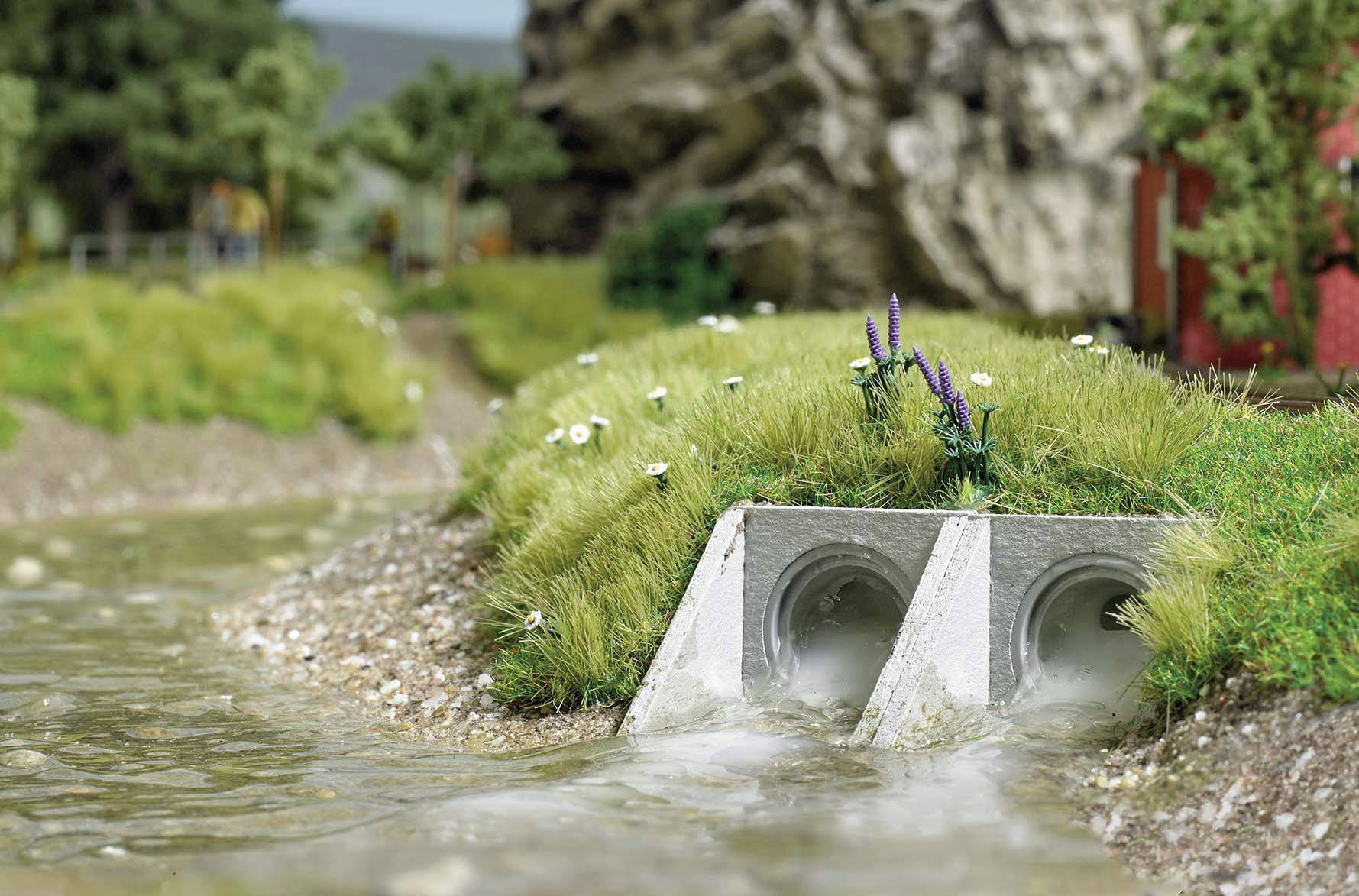 Zwei Betonrohre entwässern in einen Bach in einer ländlichen Landschaft mit Gras und Bäumen im Hintergrund.