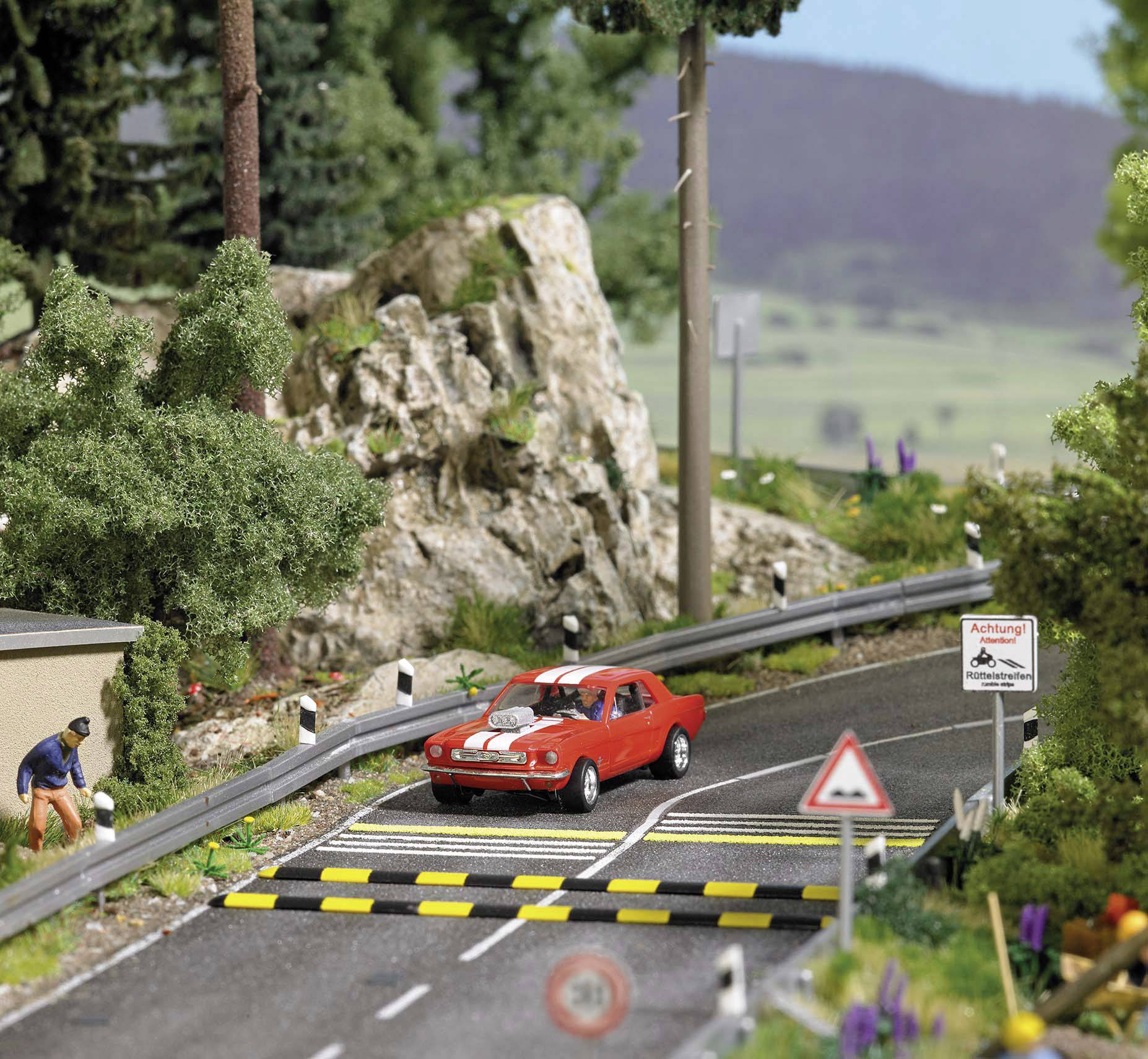 Ein rotes Auto fährt auf einer Straße mit Bodenschwelle in einer ländlichen Miniaturlandschaft. Berge und Schilder im Hintergrund.