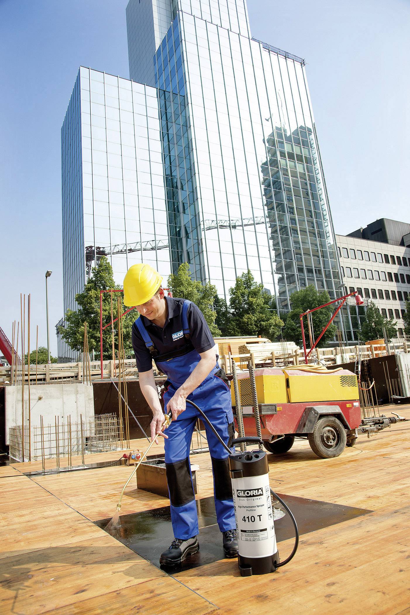 Ein Bauarbeiter mit gelbem Helm sprüht Flüssigkeit auf eine Holzplatte auf einer Baustelle vor einem modernen Hochhaus.