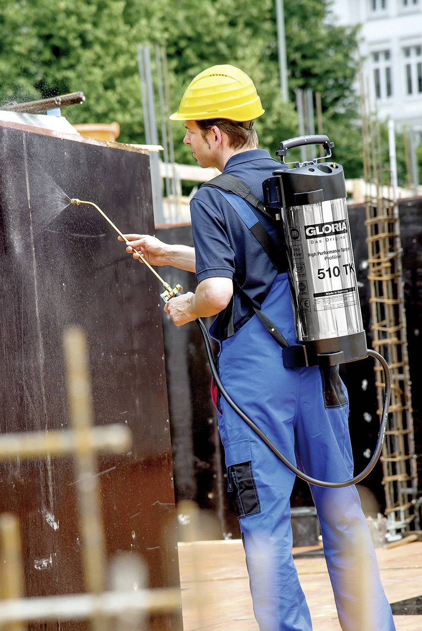 'Arbeiter sprüht mit Schutzanzug und Helm auf Baustelle schwarzen Behälter an. Trägt Rückensprüher mit Aufschrift 
