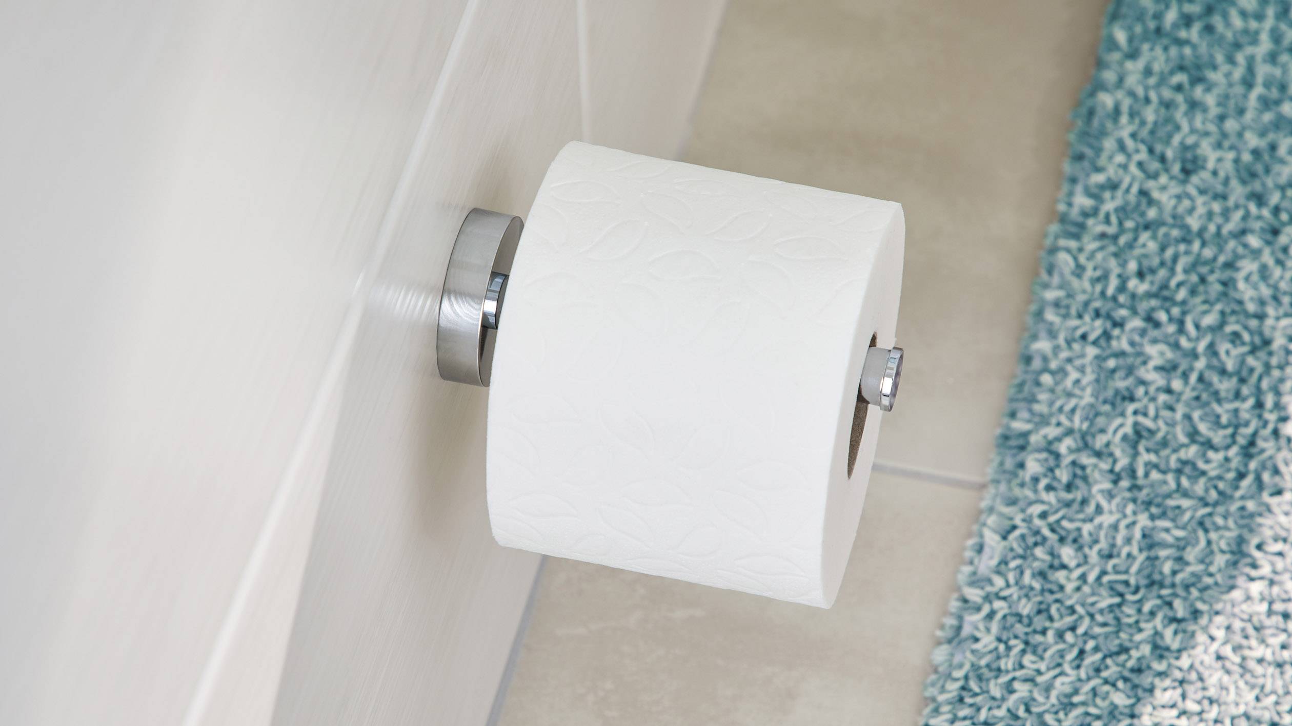 Eine Rolle Toilettenpapier an einer Wandhalterung neben einem blauen Badteppich auf Fliesenboden.