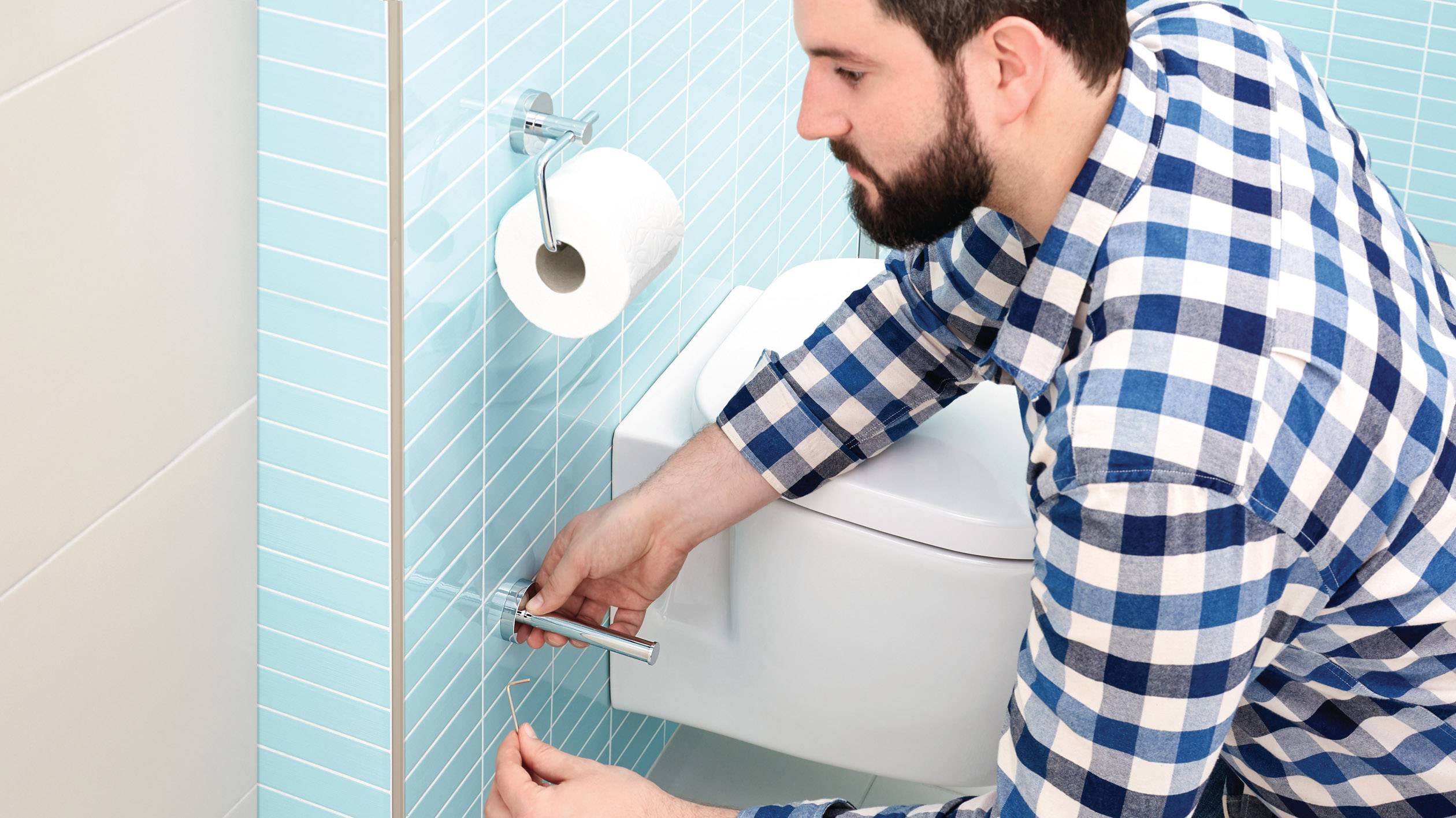 Ein Mann im karierten Hemd repariert die Halterung einer Toilettenpapierrolle im Badezimmer mit blauen Fliesen.