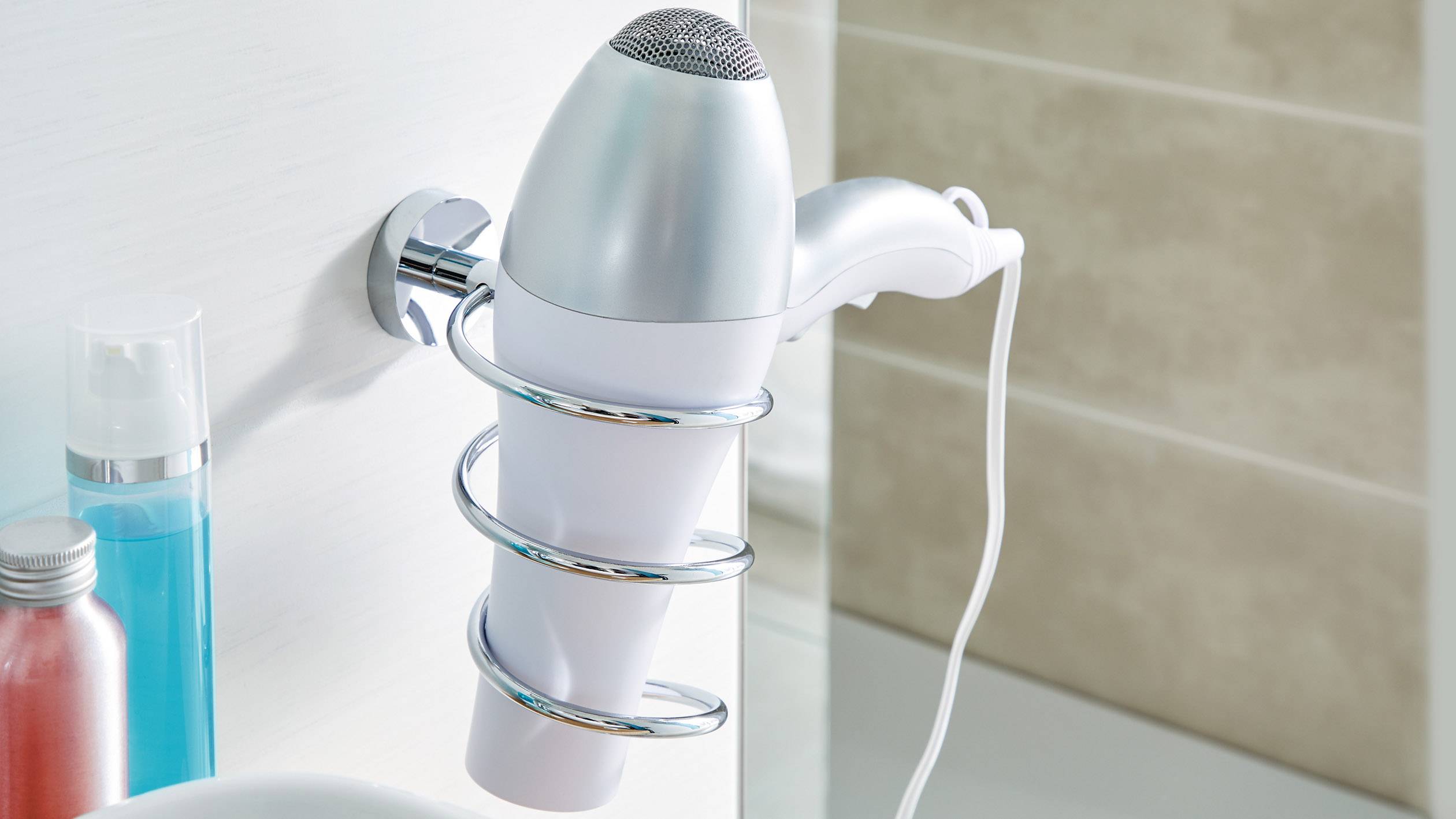 Ein weißer Haartrockner steckt in einer Wandhalterung im Badezimmer. Daneben stehen zwei Flaschen mit Pflegeprodukten.