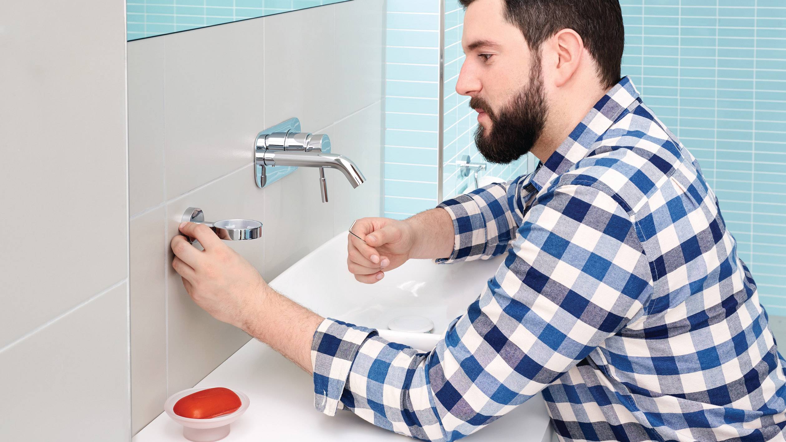 Ein Mann im karierten Hemd repariert einen Wasserhahn im Badezimmer. Auf dem Waschbecken steht eine Schale mit Werkzeugen.