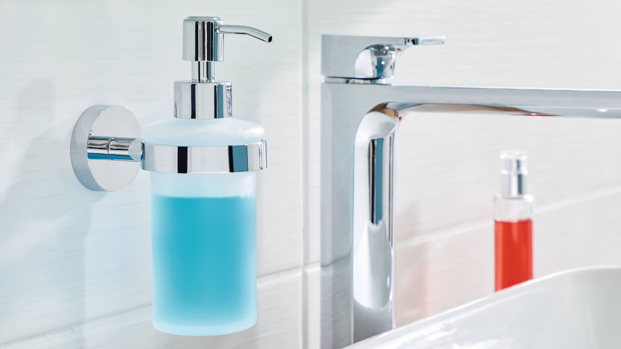 Seifenspender mit blauem Inhalt an weißer Badezimmerwand montiert, nahe einem Waschbecken mit verchromter Armatur.