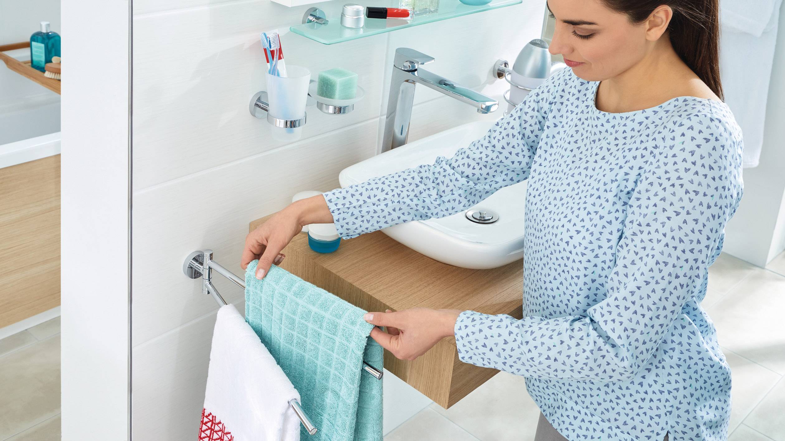 Eine Frau hängt ein blaues Handtuch an einen Handtuchhalter im Badezimmer. Im Hintergrund sind ein Waschbecken und Badaccessoires.