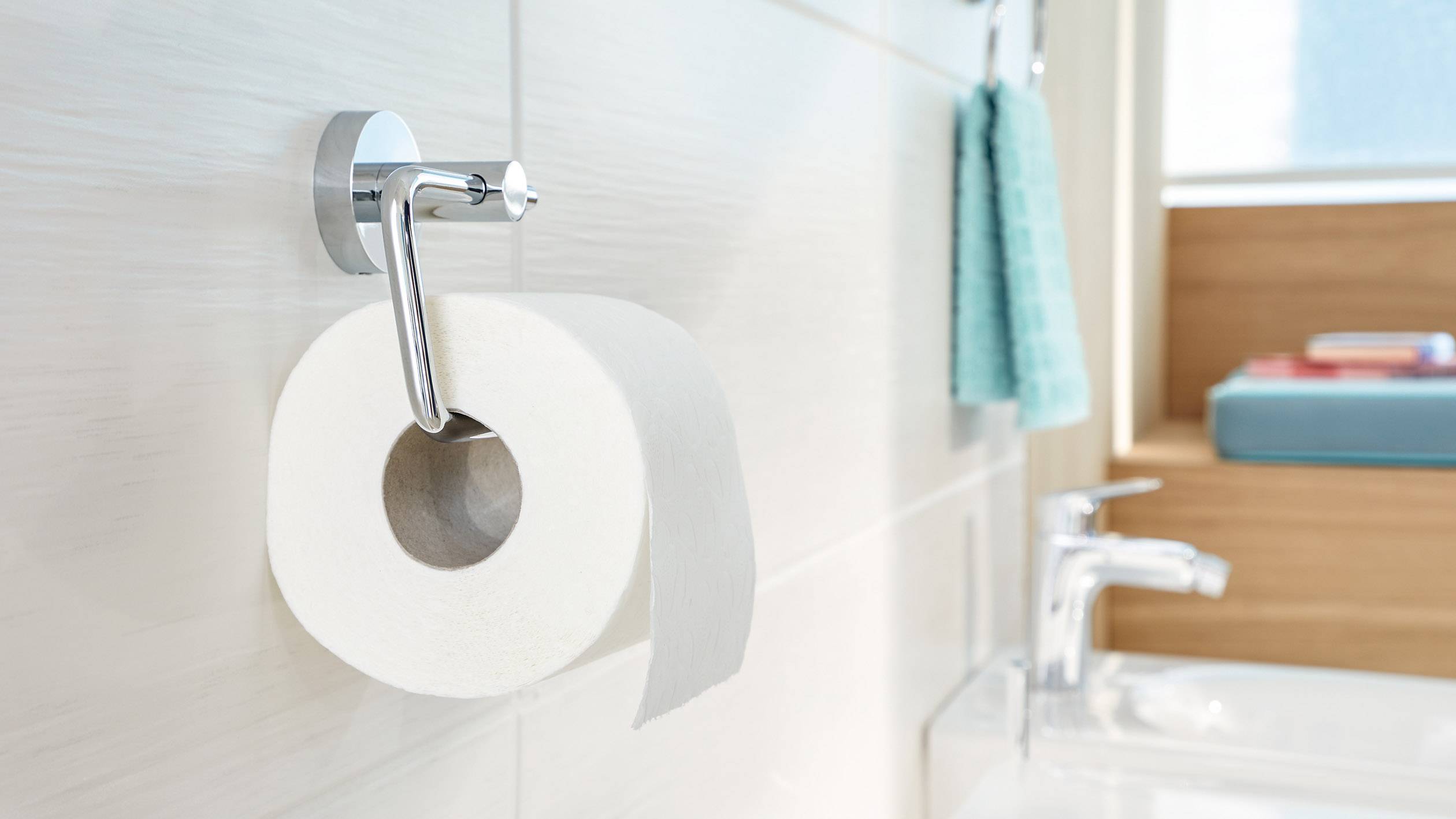 Toilettenpapierrolle hängt an einem Halter an einer weiß gefliesten Wand neben einem Waschbecken. Ein türkisfarbenes Handtuch ist sichtbar.