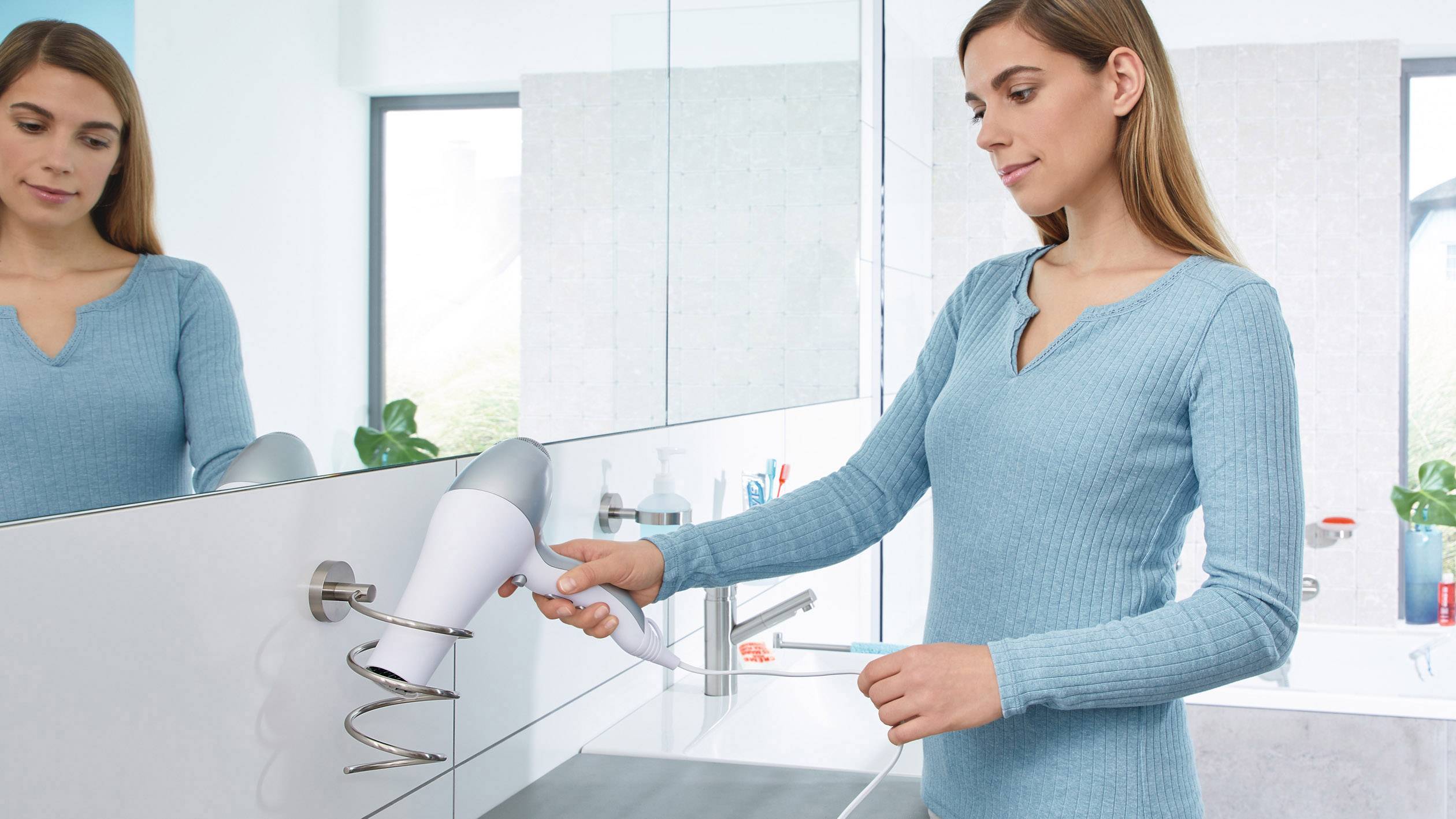 Eine Frau im blauen Pullover legt einen Haartrockner in eine Wandhalterung im Badezimmer.